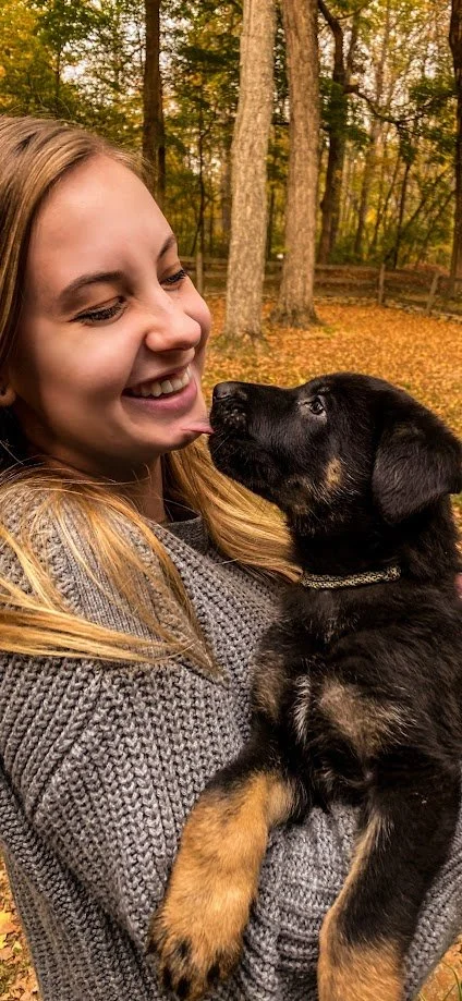 Trainer Sara smiling and holding a German Shepherd puppy that is looking up to her and licking her face sweetly.