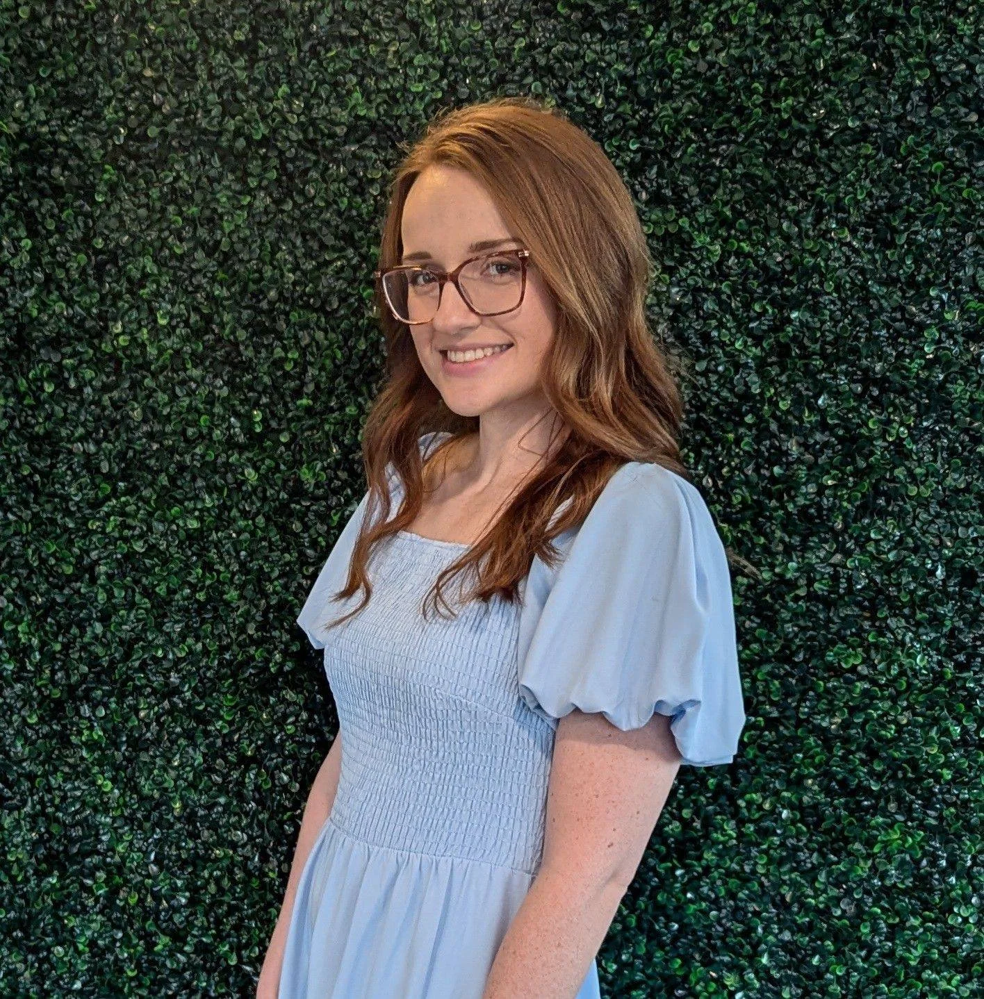 Red haired woman in a blue dress against a green foliage background.