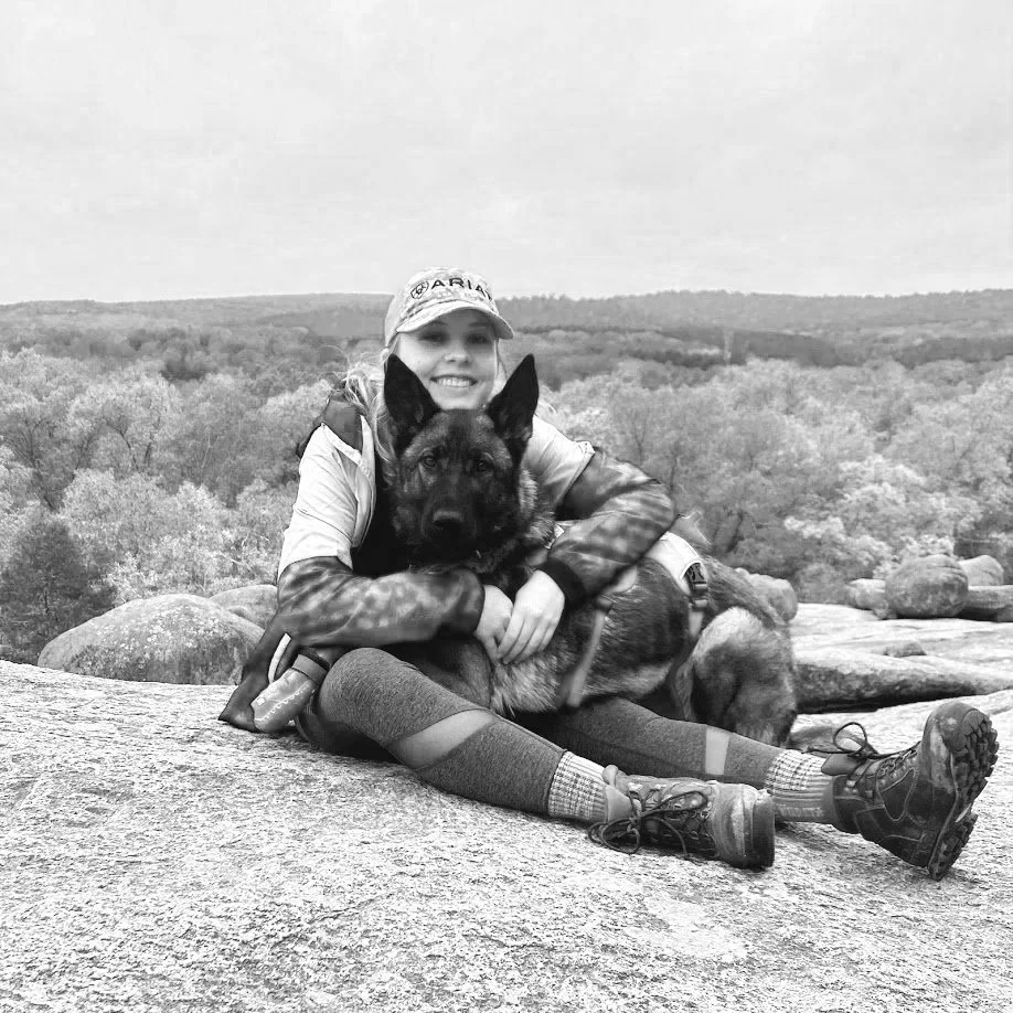 A woman with a hat and outdoor gear sitting on a rock, hugging a German Shepherd dog against a landscape of trees and rocks.