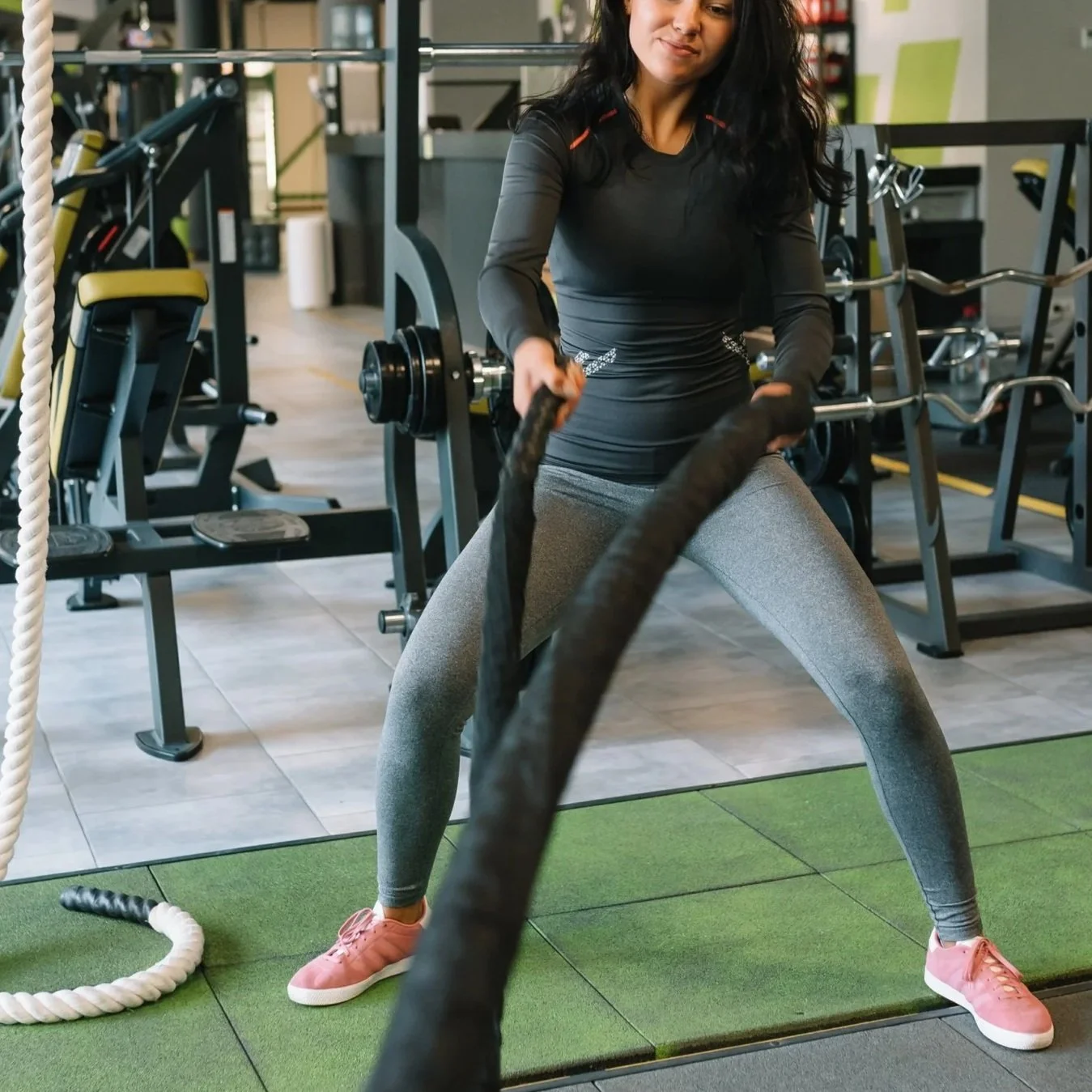 A woman in workout clothes is exercising with battle ropes in a gym, surrounded by workout equipment and a green exercise mat.