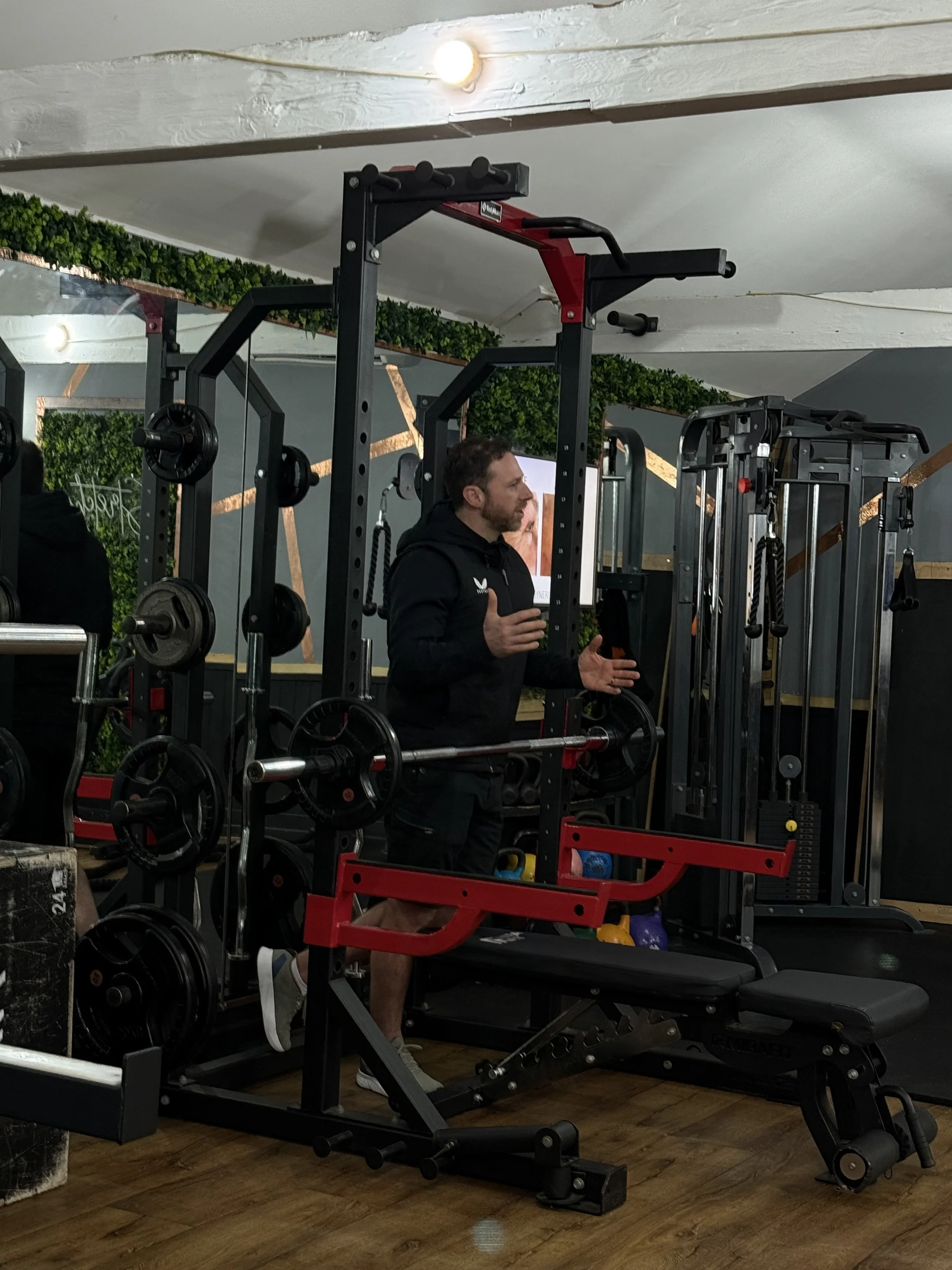 A man with a beard wearing a black hoodie talking and gesturing with his hands in a gym, surrounded by gym equipment including weight plates and resistance machines.