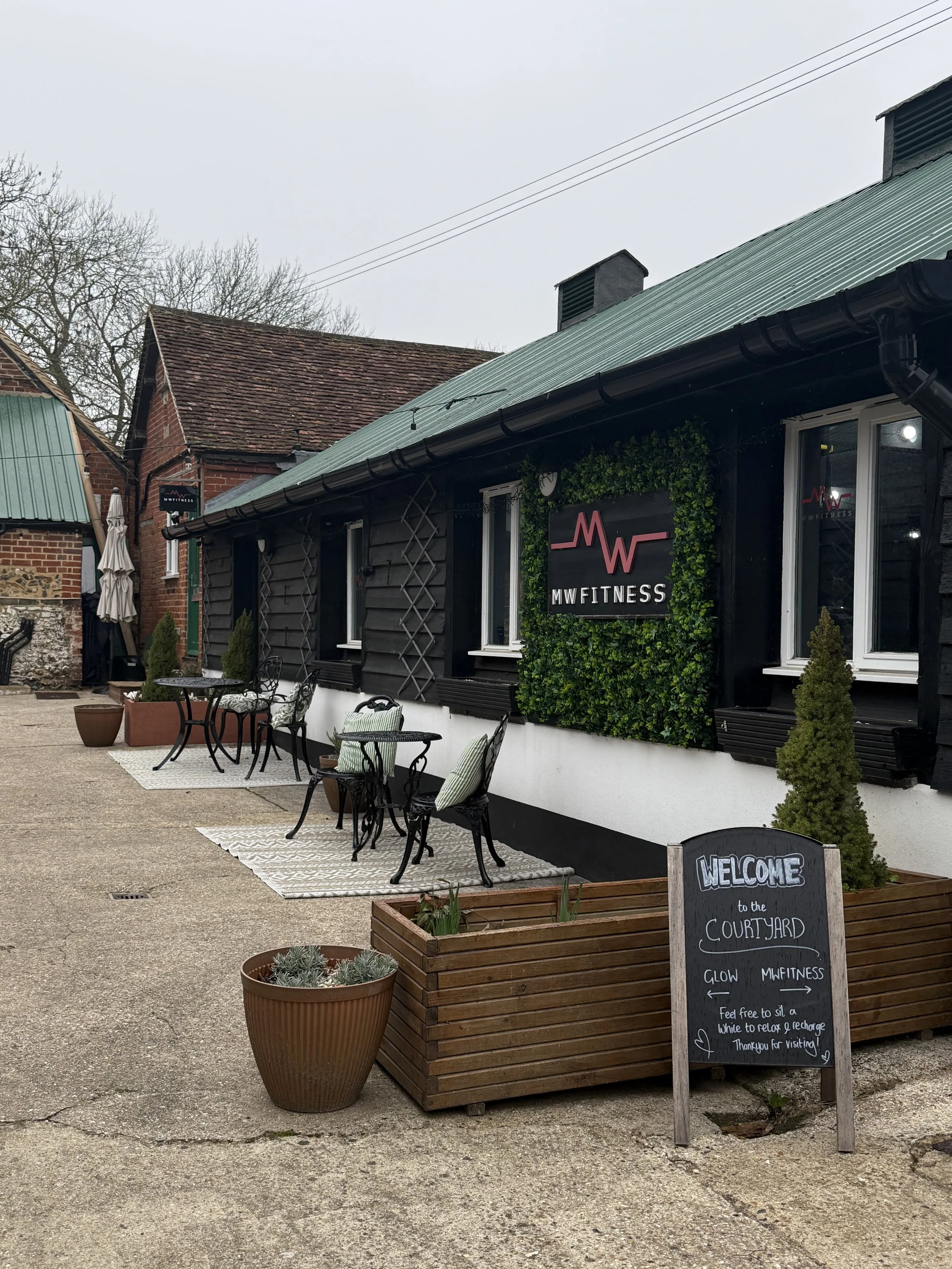 Exterior of a fitness studio named 'MW Fitness' with outdoor seating and decorative plants. A chalkboard sign welcomes visitors to the courtyard with amenities like glow and MW Fitness, and urges customers to relax and recharge.