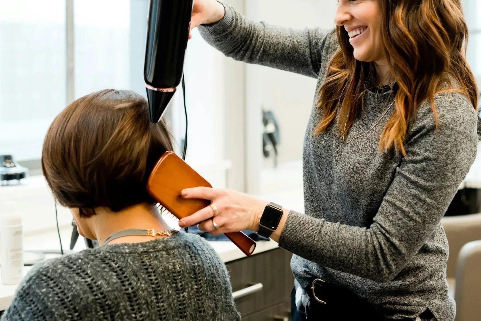 A woman is blow-drying and styling another woman's hair in a salon. The woman receiving hair styling has short brown hair and is holding a hairbrush, while the stylist is smiling and holding a hairdryer.