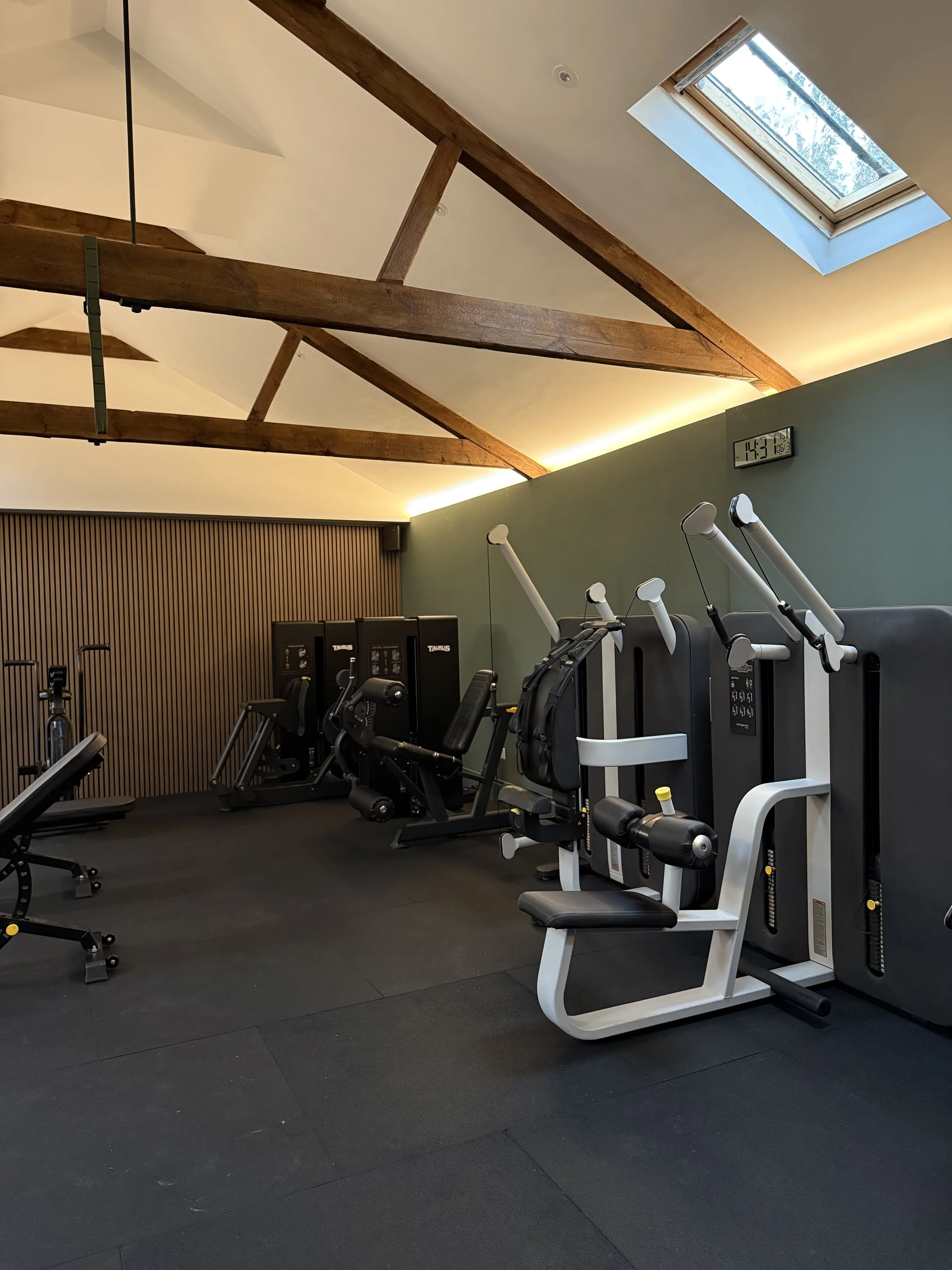Gym with various exercise machines, wooden ceiling beams, a skylight window, and a digital clock on the wall.