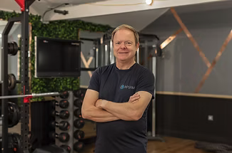 A man standing in a gym with arms crossed, wearing a black t-shirt, with gym equipment and a TV in the background.