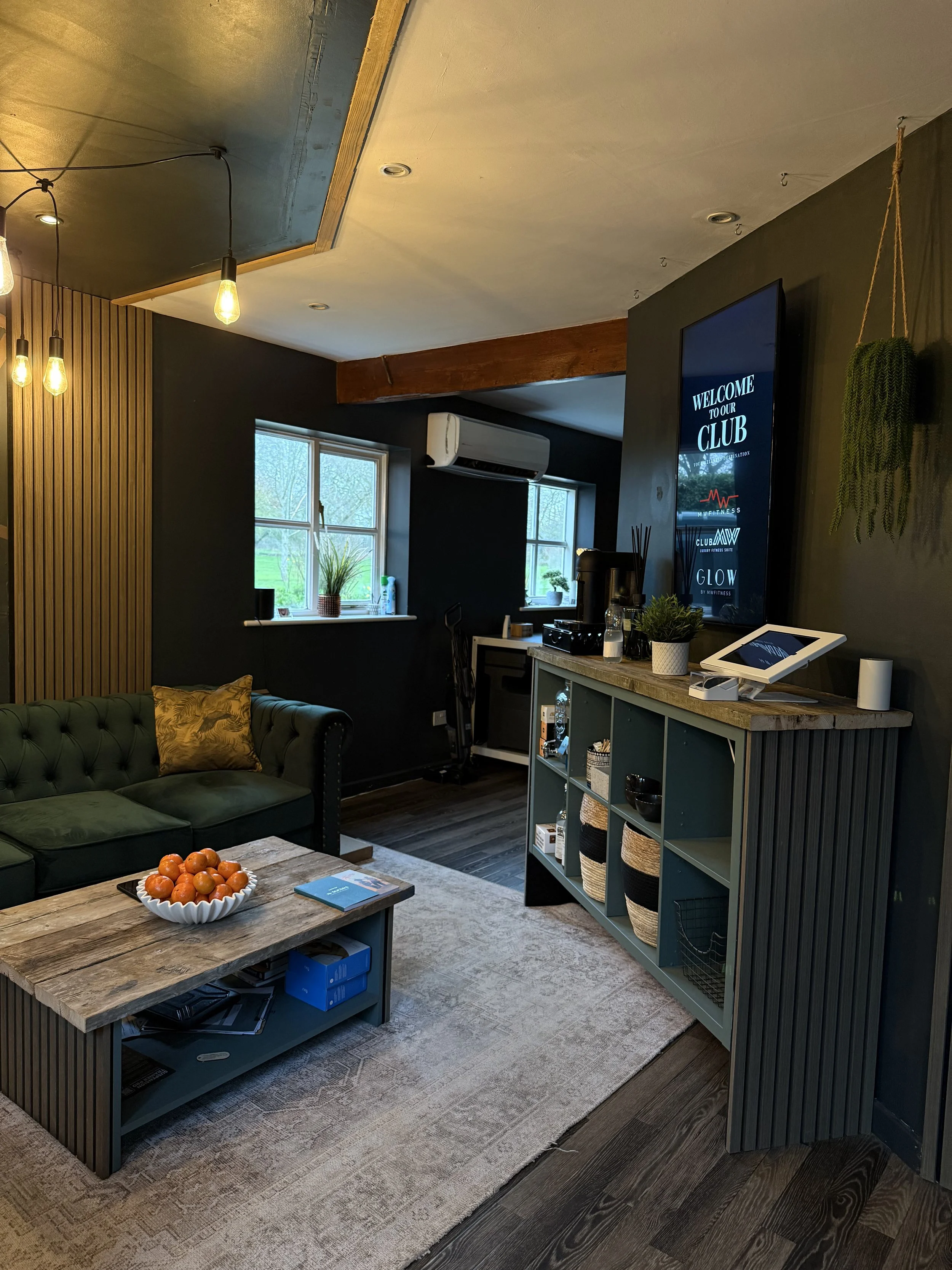 A cozy living room with a green tufted sofa, a rustic wooden coffee table with cherries, and a beige patterned rug. There are large windows with green plants, a dark accent wall with a TV displaying a welcome message, and stylish lighting fixtures. A teal shelving unit holds baskets and decor, and a small bar area is visible.