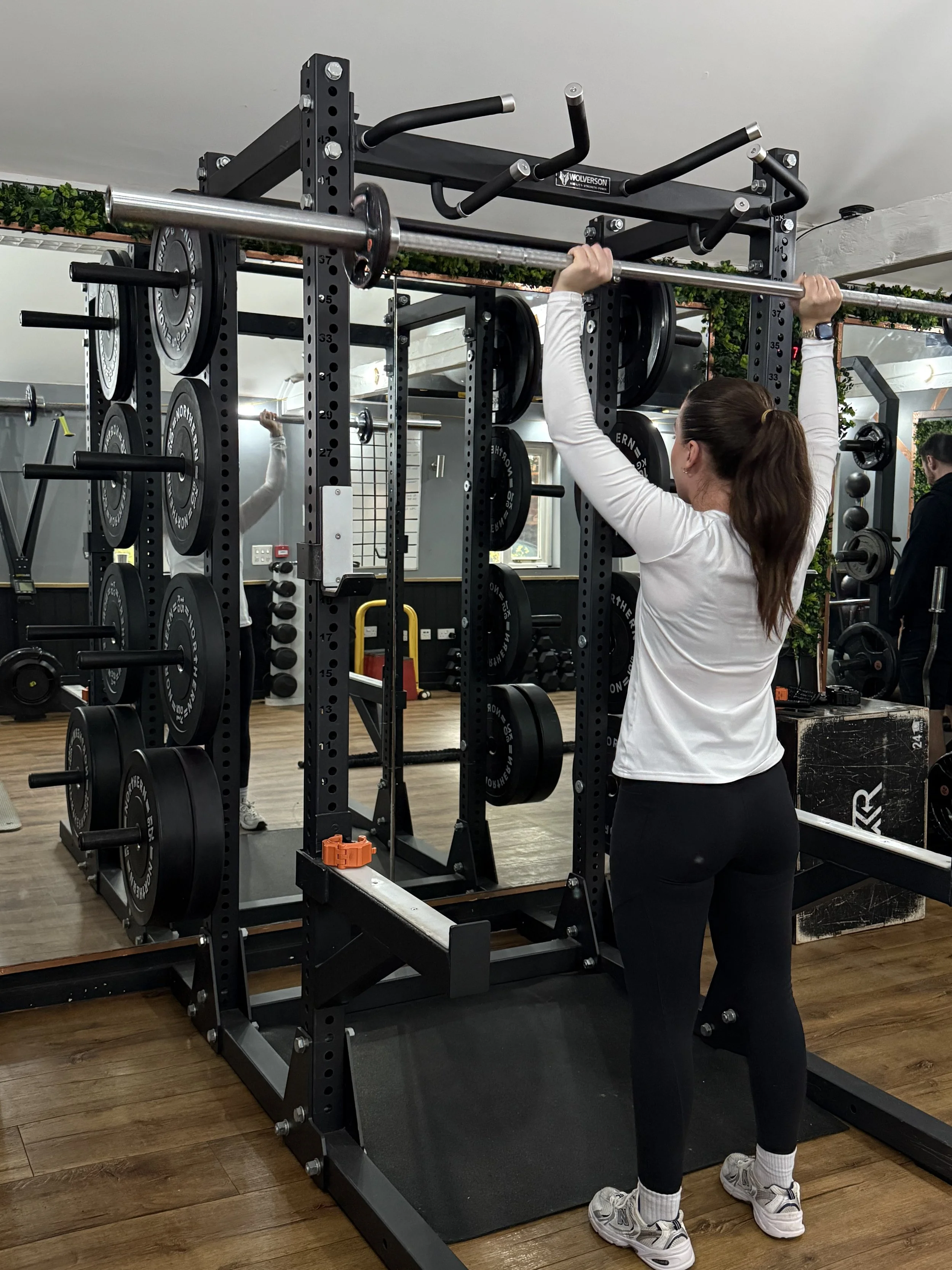 A woman lifting a barbell in a gym. She is wearing a white long-sleeve shirt, black leggings, and sneakers, with her hair in a ponytail. The gym has various weights and equipment, with mirrors and a wooden floor.