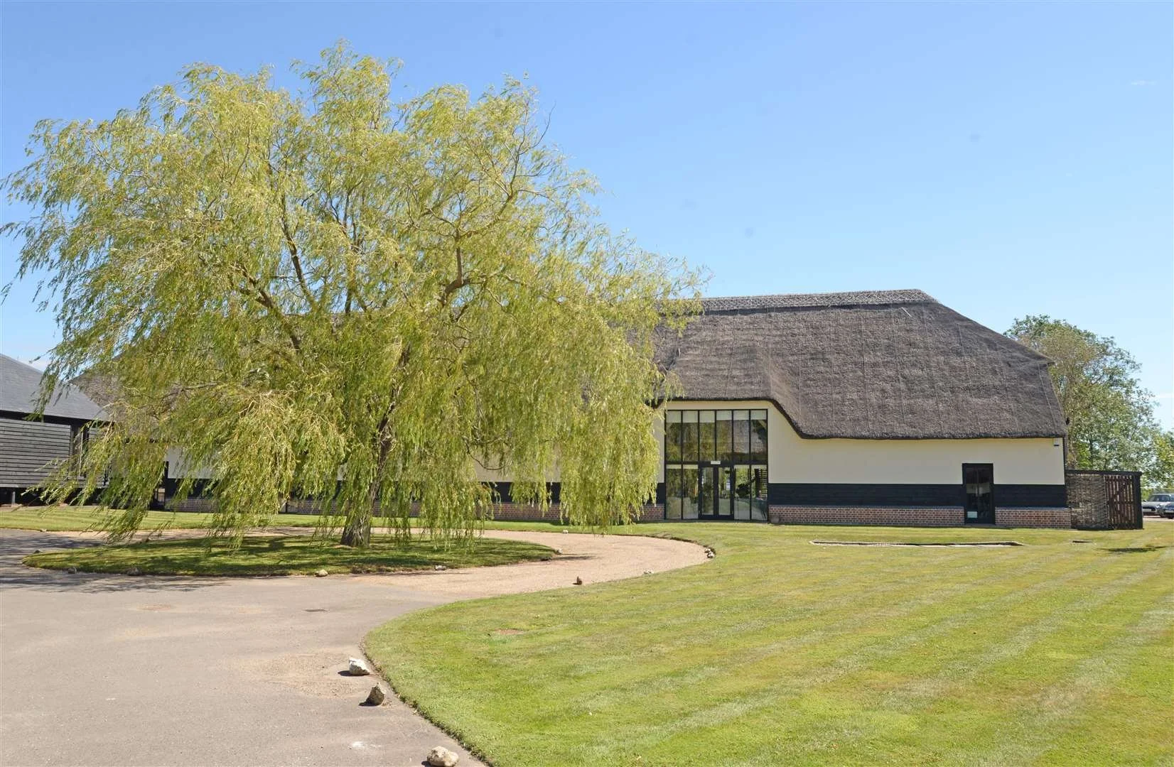 A large building with a thatched roof and white walls with black trim, set beside a large green tree and a well-maintained grassy area, under a clear blue sky.