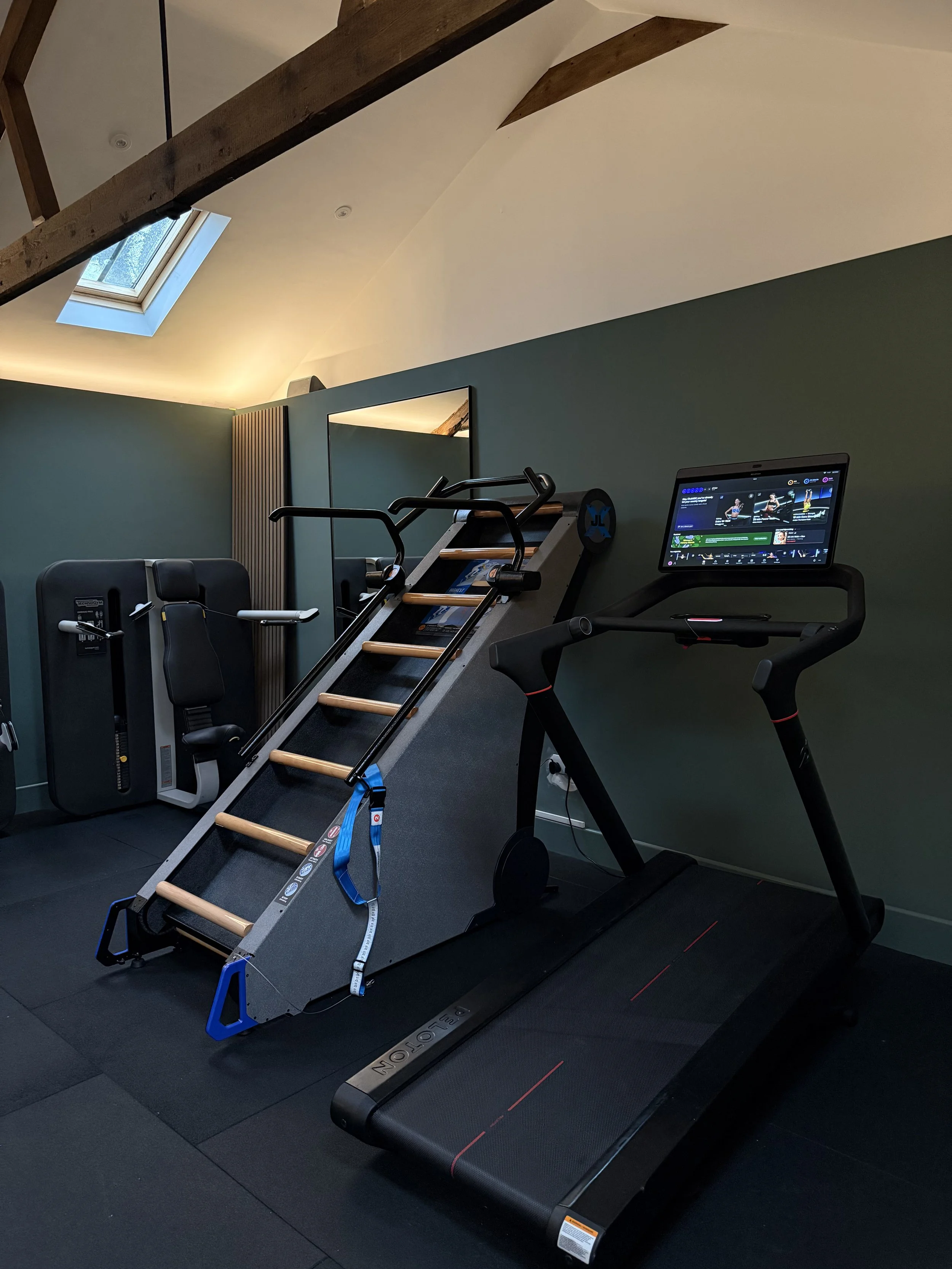 Indoor gym with a treadmill, rowing machine, and stair climber on black flooring. There is a green wall, a mirror, a window in the ceiling, and wooden beams.