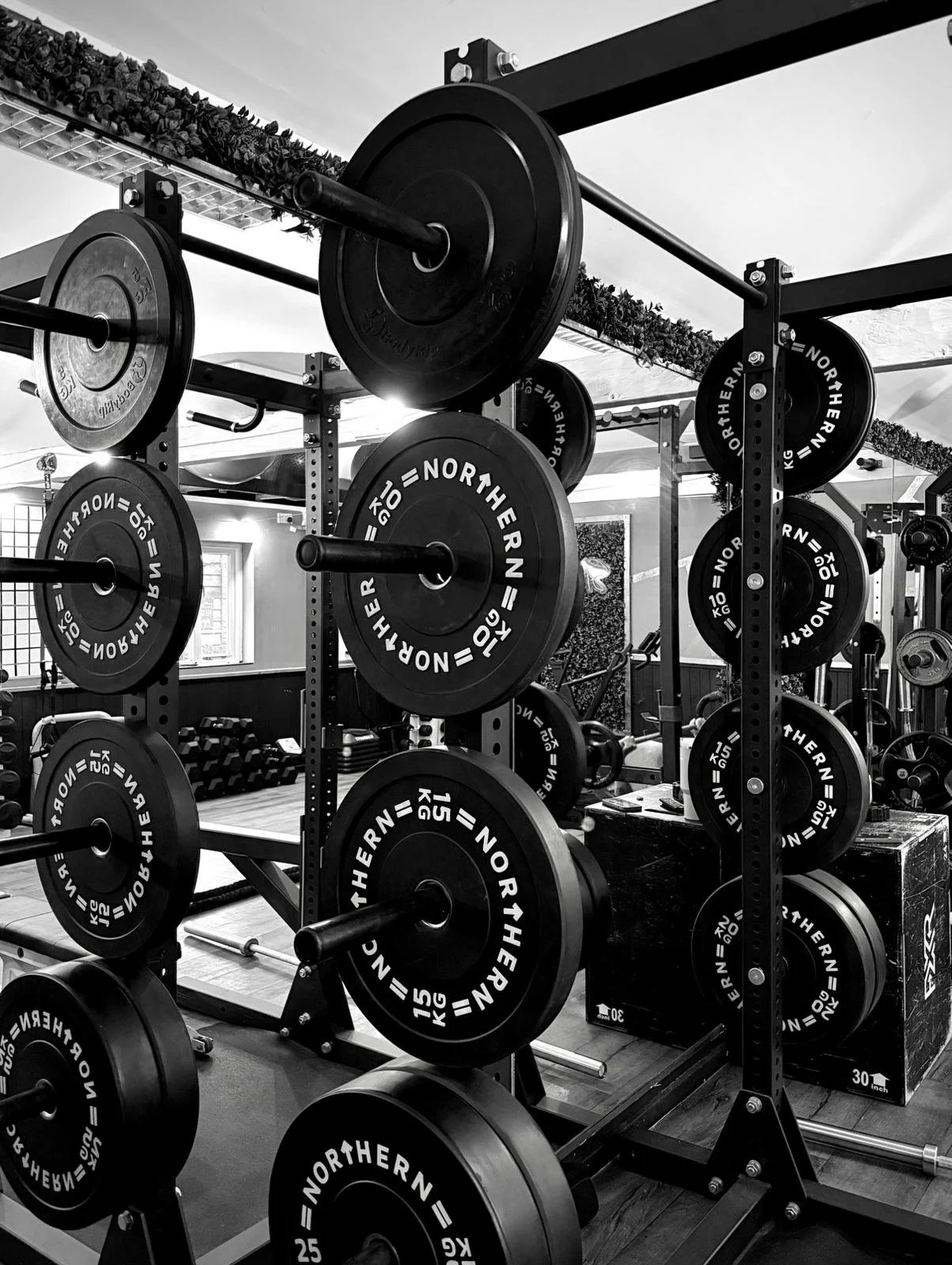 A black-and-white photo of a gym with weight racks filled with various weight plates, barbells, and equipment for strength training.