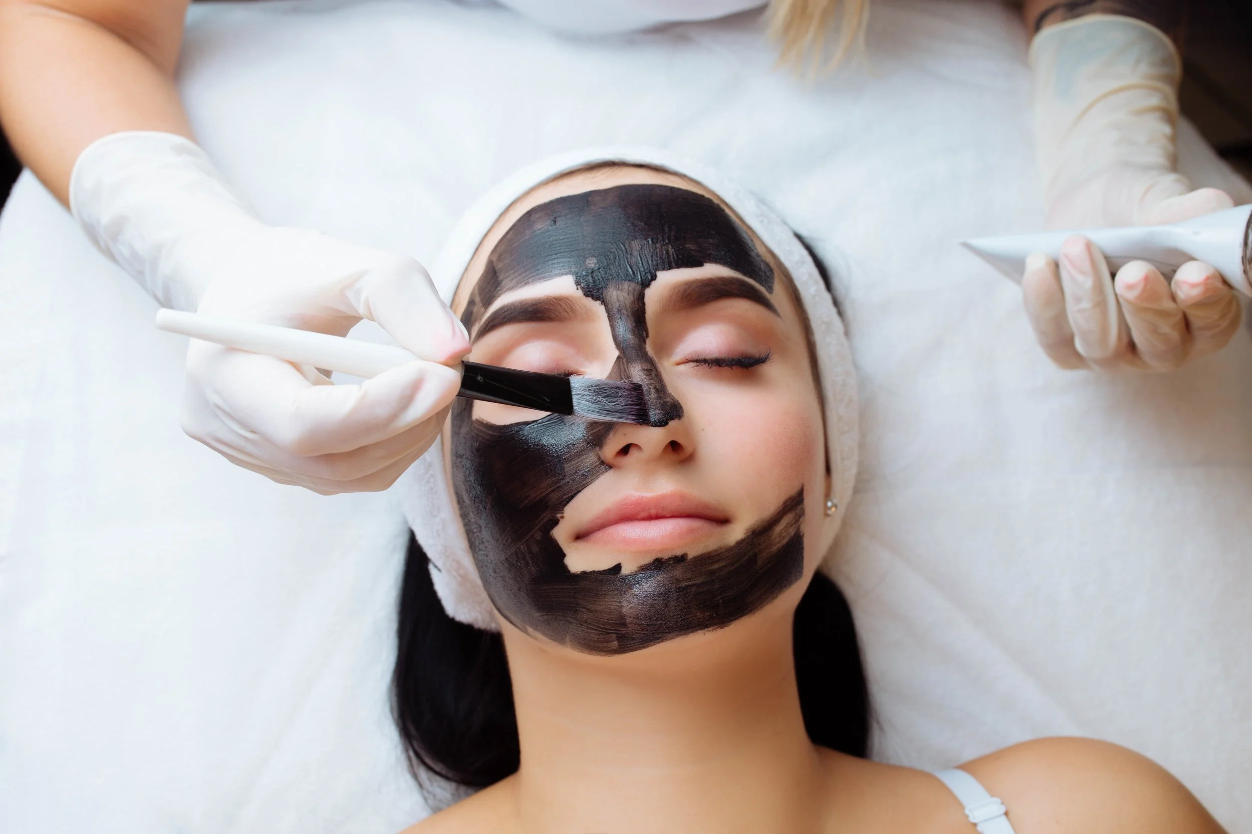 A woman is lying down with her eyes closed, getting a facial treatment. She has a dark facial mask being applied to her face by a skincare professional wearing gloves.