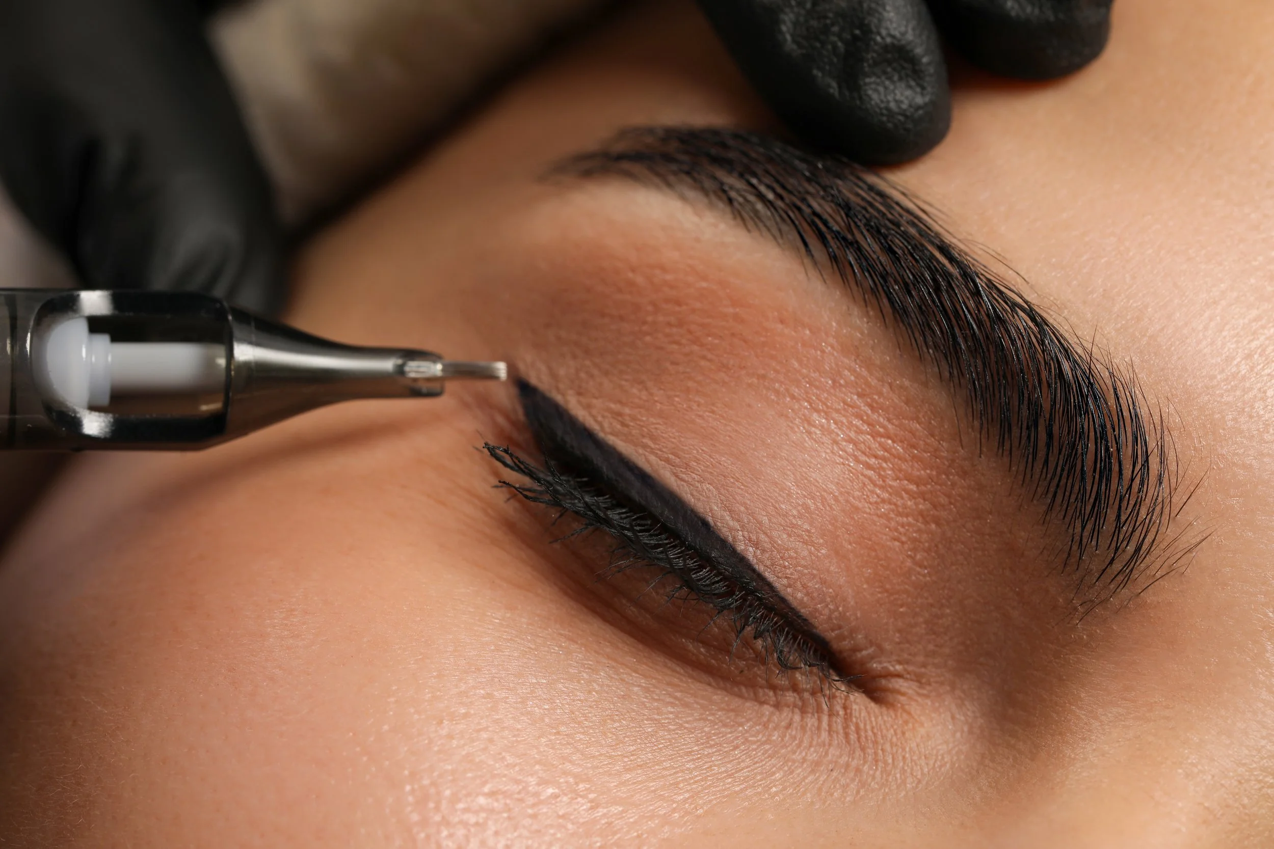 Close-up of a person getting black eyeliner applied to their upper eyelid with a fine-tipped pen, while another hand in a black glove holds the eyelid steady.