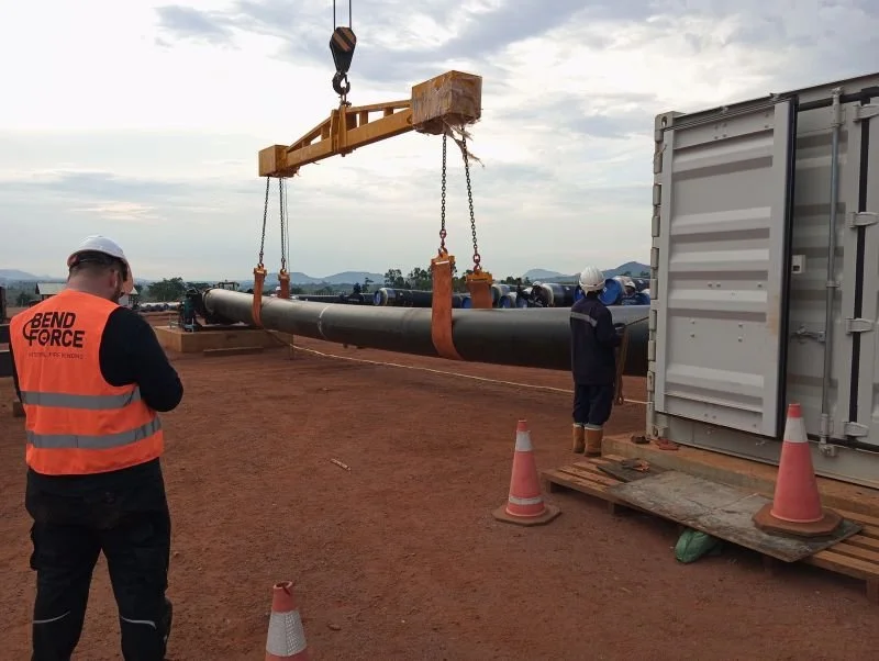 Workers on a construction site guiding a large pipe using a crane with a hook. One worker wears an orange safety vest with 'BEND FORCE' printed on the back, and another stands near a shipping container. Orange traffic cones mark the area.