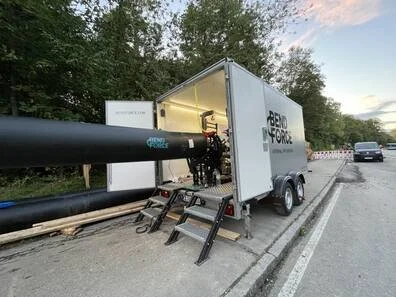 A mobile underground utility inspection vehicle with a large black pipe extending from it, parked on the side of a road with trees and cars in the background.