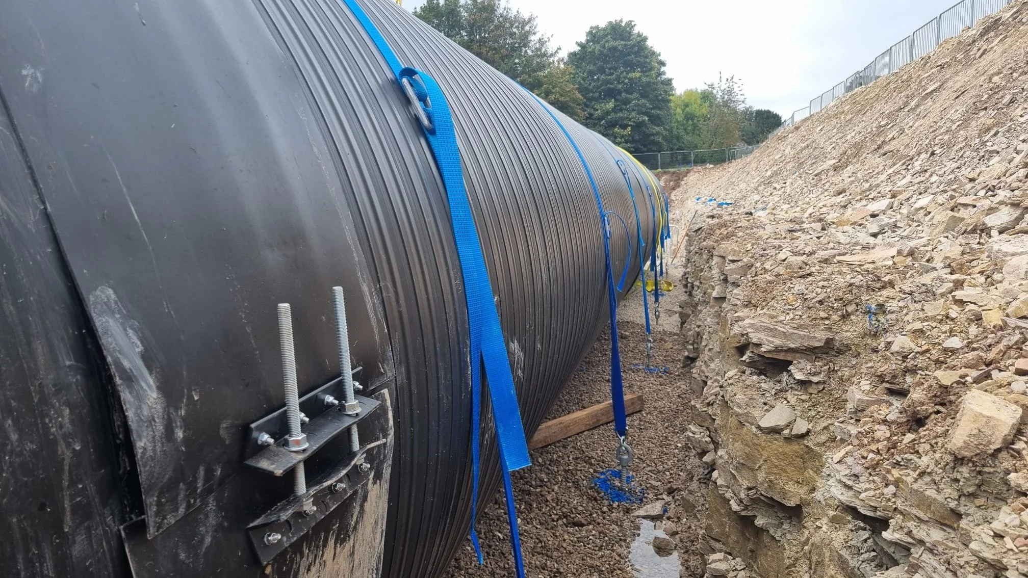 Large black plastic underground water or oil pipeline secured with blue straps, running parallel to a dirt and rocky excavation site with a metal fence on top of the embankment in the background.