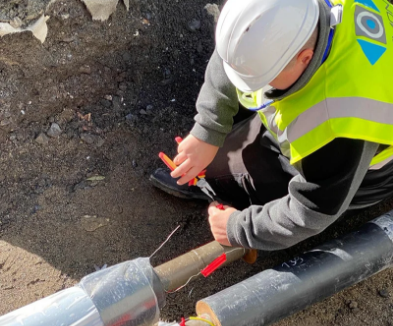 A construction worker in a neon safety vest and white helmet working on underground piping, using a tool to connect pipes.