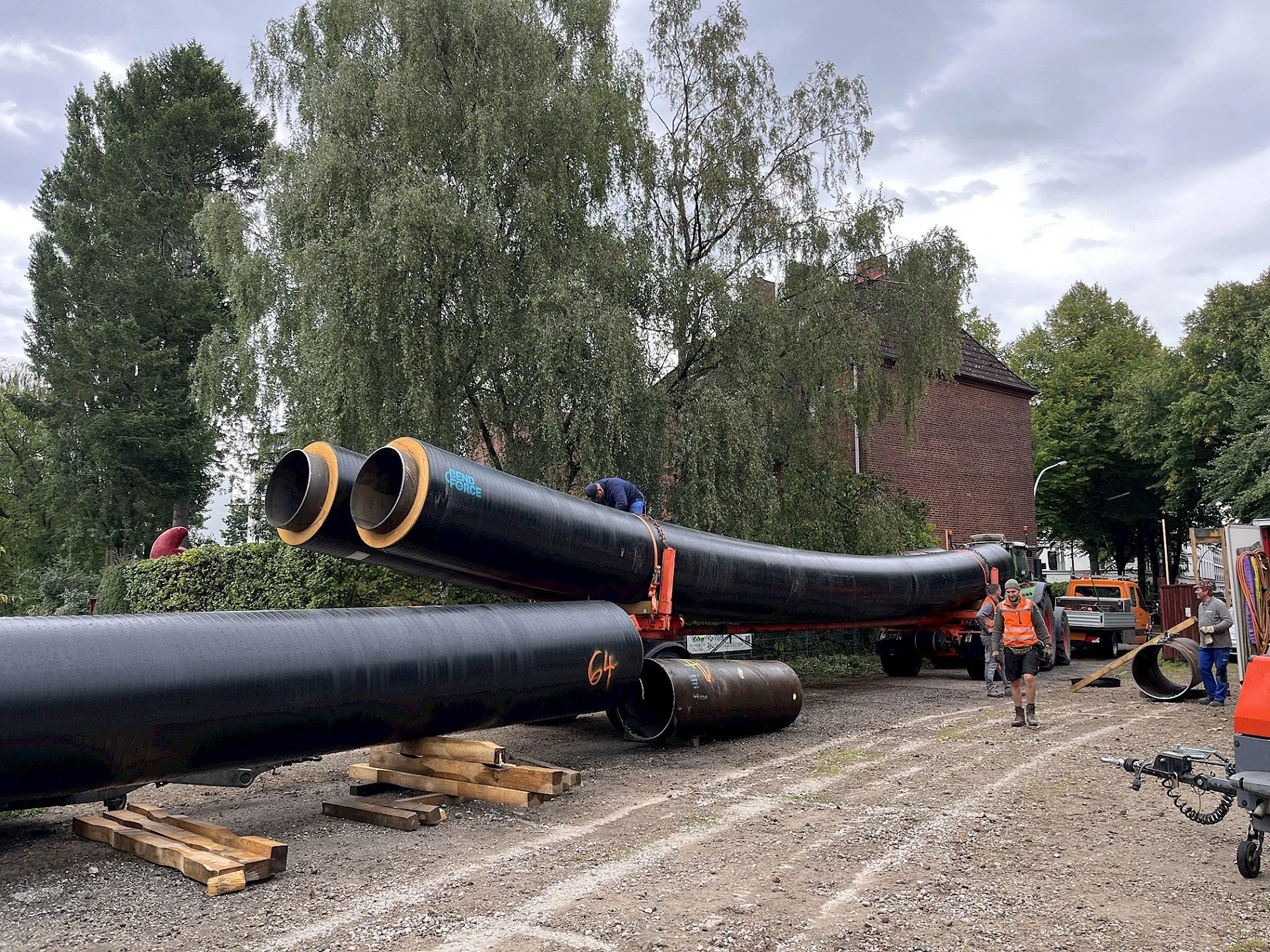 Workers setting up large black pipes on a construction site. The pipes are resting on wooden supports, and there are trees and a brick building in the background. One worker is on top of the pipe, while others are on the ground.