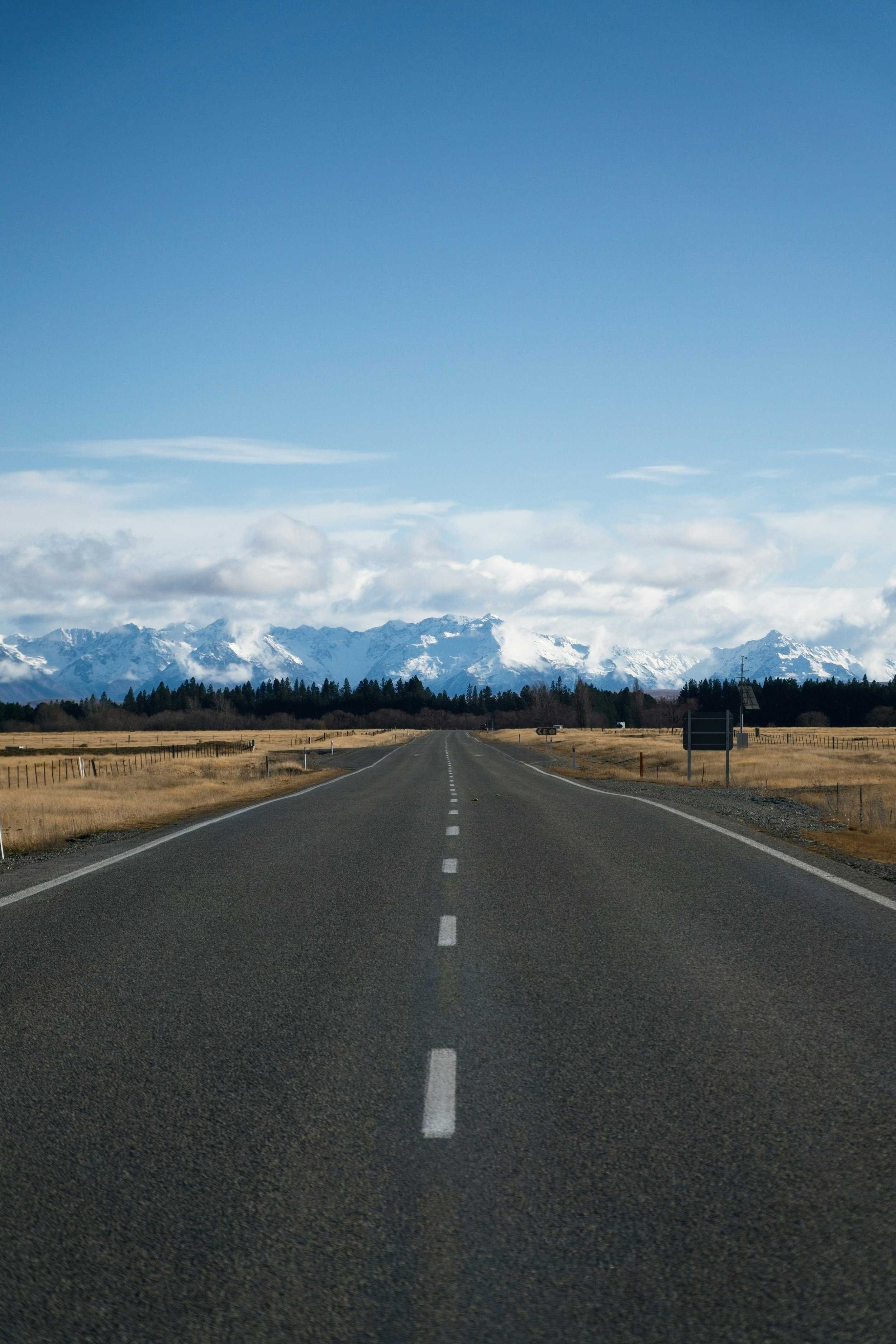 Open road leading towards snowy mountains under a blue sky with clouds.