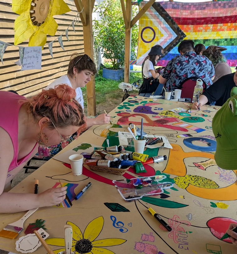 People working on collaborative artwork around a large table. There's a big pride flag in the back with another group of people huddled together. Paint and brushes are scattered around the middle of the table.