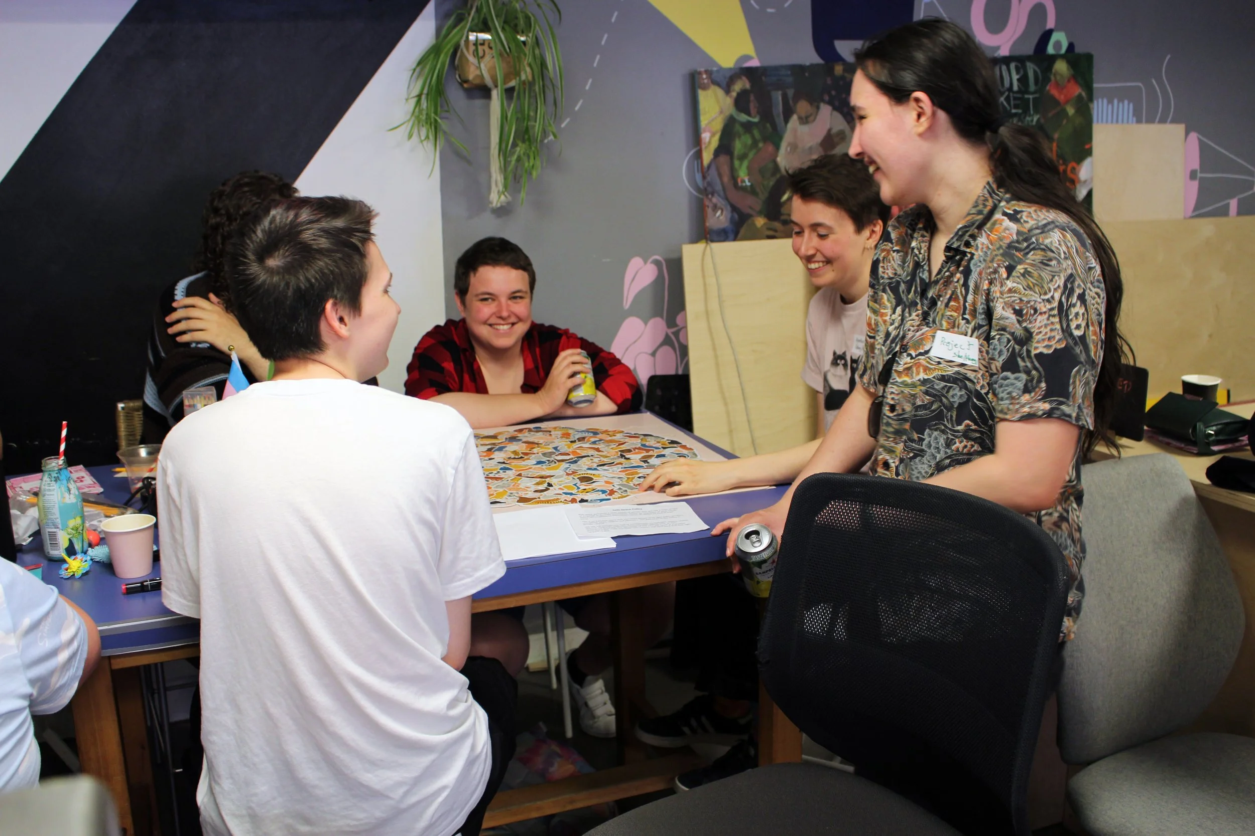 Four people around a large table, completing an activity together and smiling. They have name & pronoun badges on their clothes.