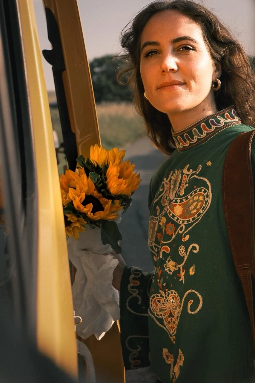 A woman with brown wavy hair holding a bouquet of sunflowers stands outside near a yellow vehicle, during golden hour.