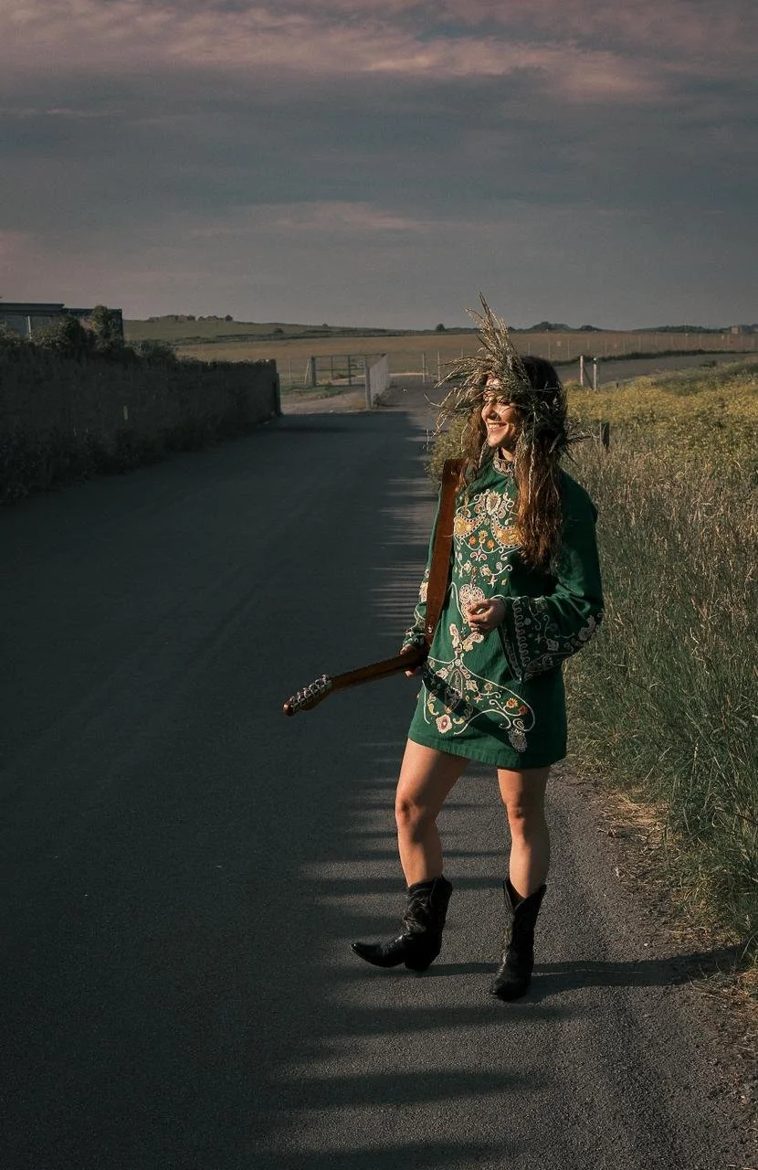 A woman standing on a rural road holding an electric guitar, wearing a green embroidered dress, black boots, and a fringe hat, with the sun setting in the background.