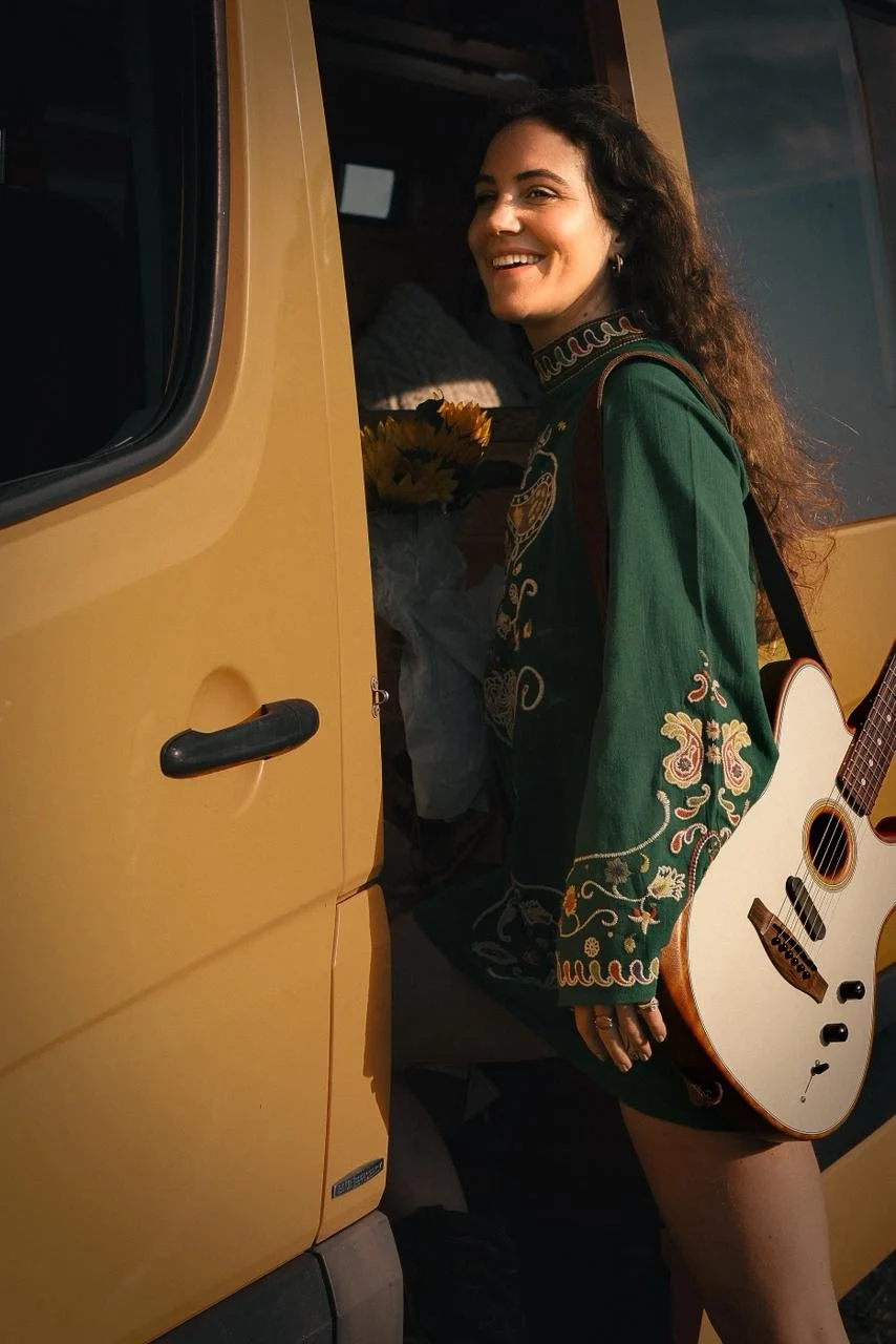 A woman with long curly hair smiling and holding a guitar, standing next to a yellow van with its door open. She is wearing a green embroidered jacket and is holding a bouquet of yellow flowers.