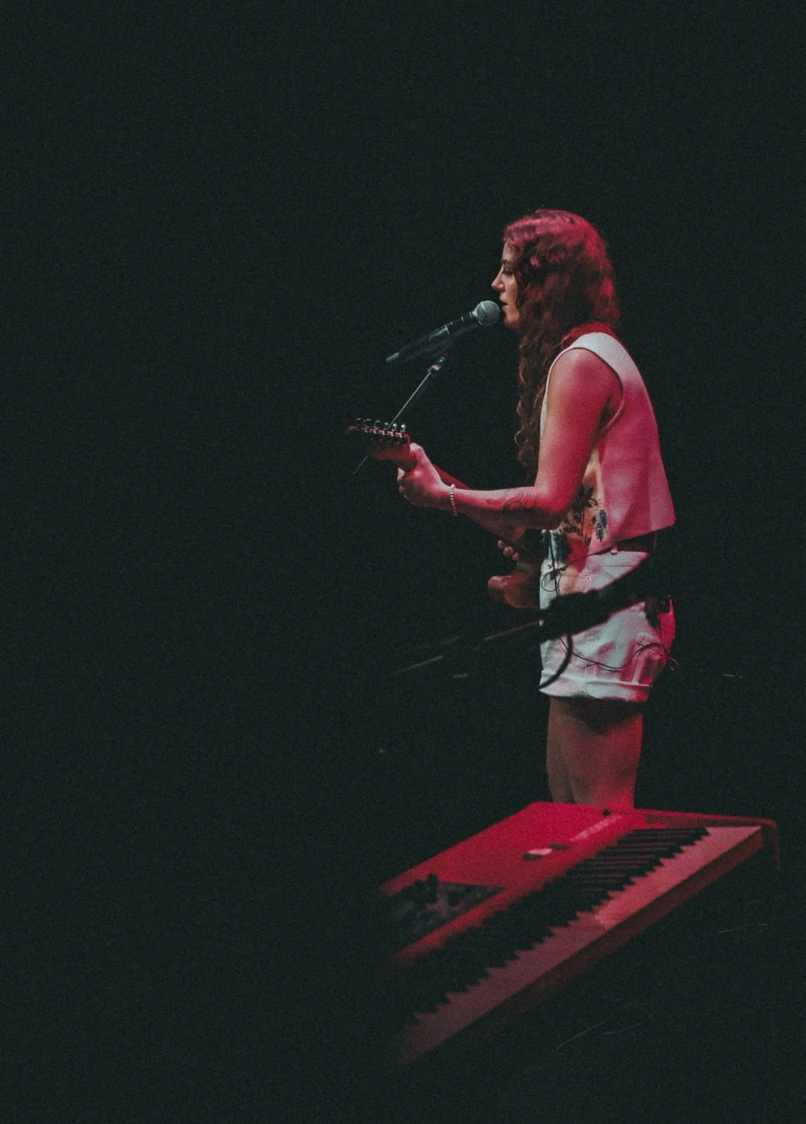 A woman with curly hair, wearing a sleeveless top and shorts, singing into a microphone on stage with a keyboard in front of her.