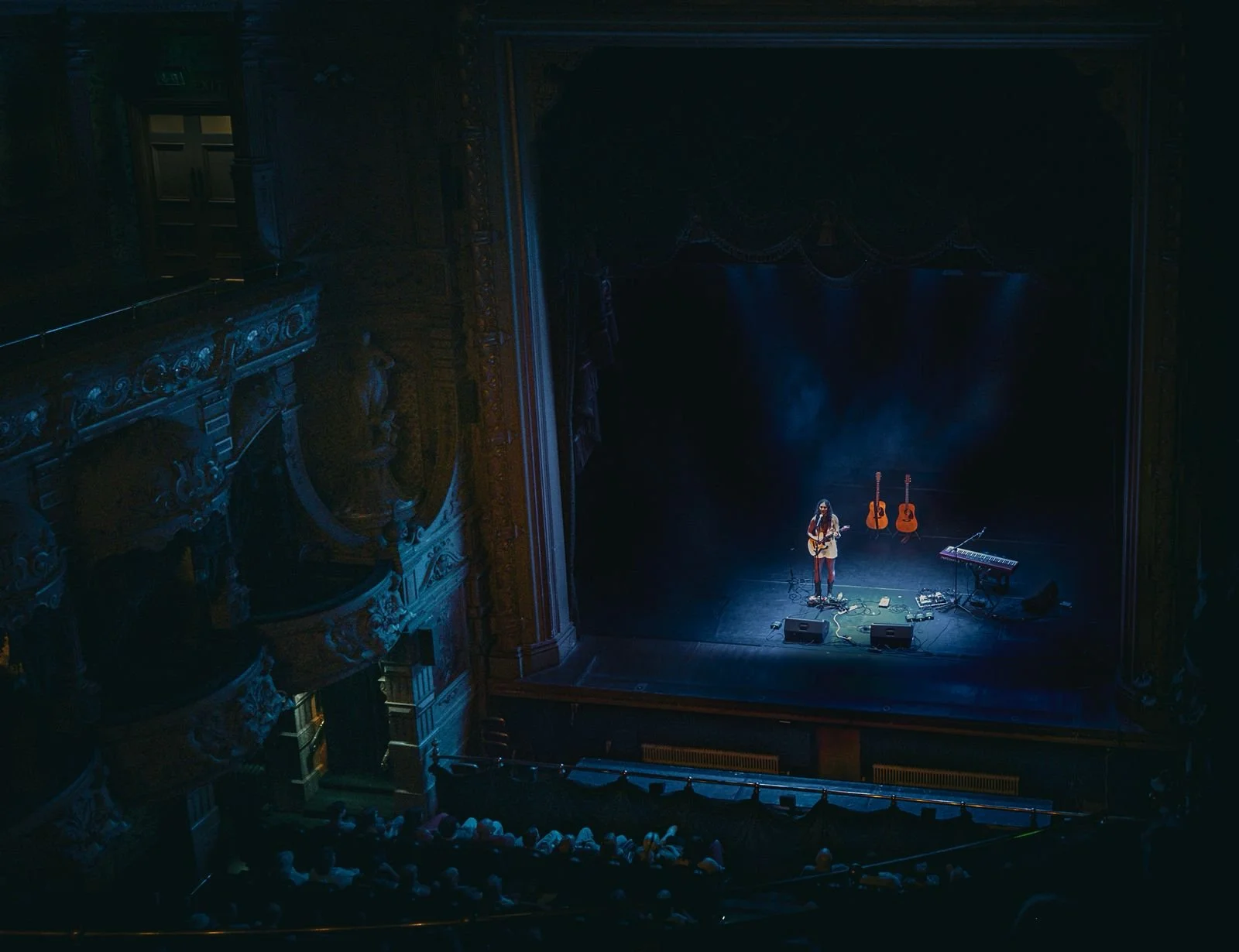A solo musician with long hair playing an acoustic guitar on a darkened theater stage, with two additional guitars and a keyboard behind them, and audience members seated in front.