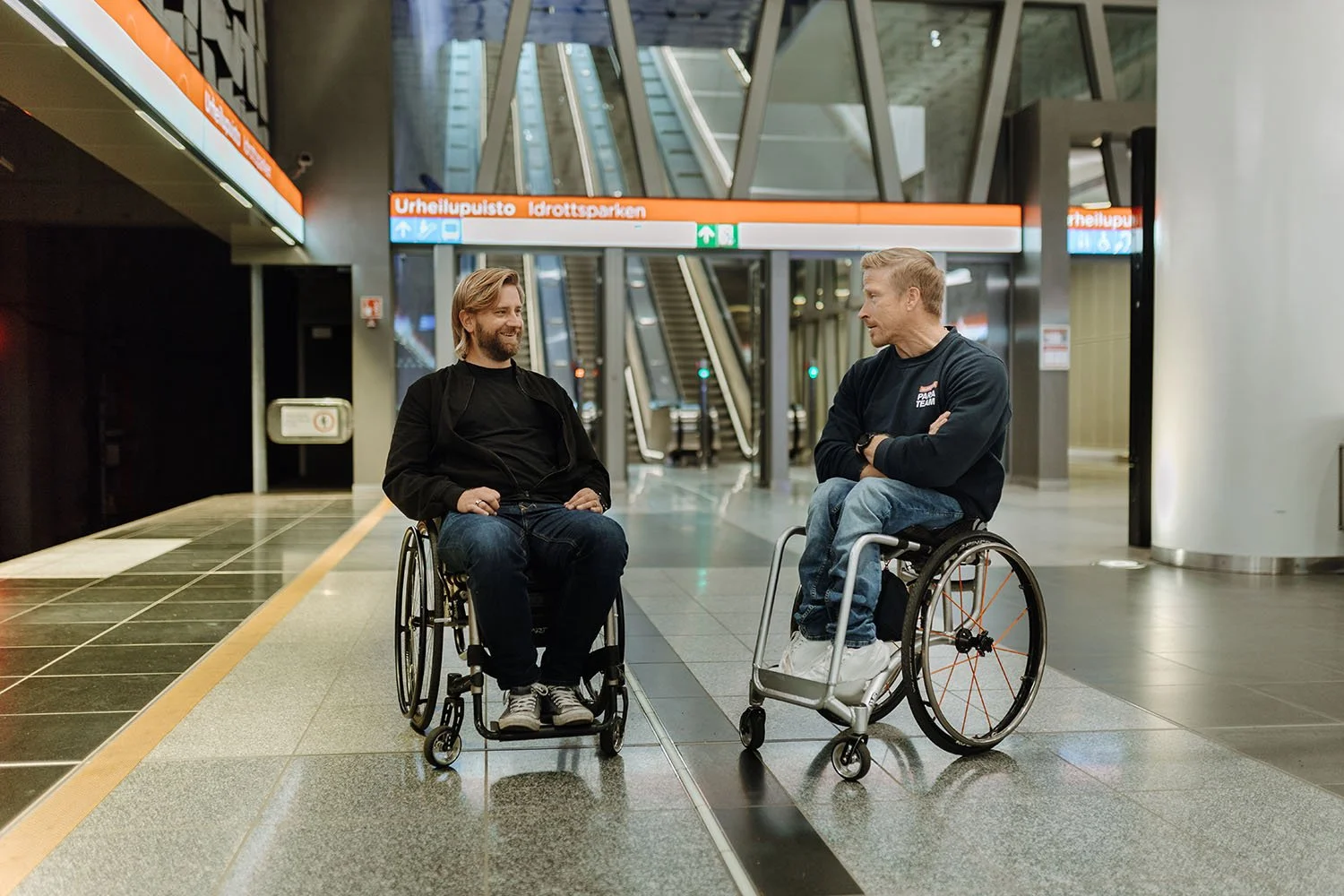 Kaksi miestä istuu pyörätuoleissa keskellä metroasemaa, keskustellen ja hymyillen. Taustalla visiirimaiset portaat ja digitalinen opastusnäyttö.