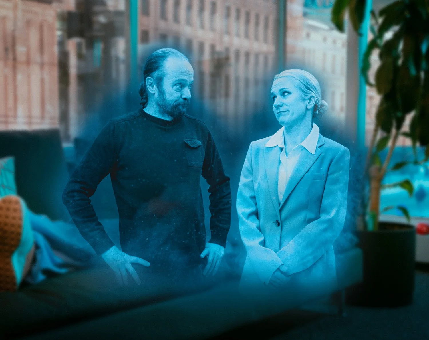 Kaksi ihmistä keskustelevat ikkunan edessä, toinen on mies ja toinen nainen. Heidän taustansa on kaupungin rakennuksia ja ikkunassa näkyy ulkopuoli.