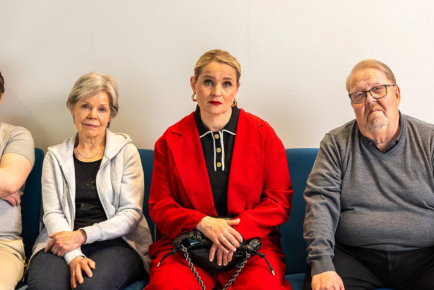 Kolme aikuista istuu penkillä vastaanotossa, yksi keskellä nainen punaisessa takissa, toiset kaksi nuorempia ja vanhempia.