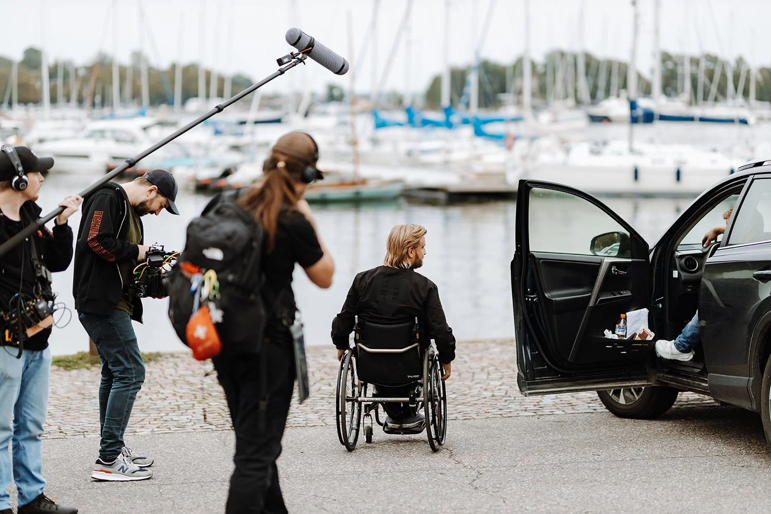 Filmi- tai televisiolähetyksen kuva moran rannalla, jossa ihmisiä, mukaan lukien mies pyörätuolissa ja ohjaaja, jotka debatoivat ja kuvaavat, ja auto parkkeerattuna.