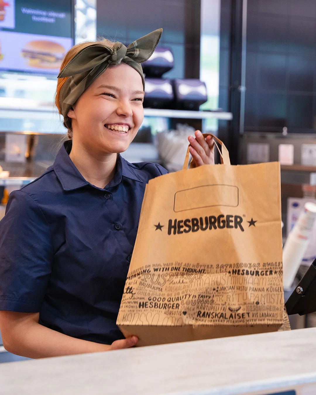 Hymyilevä nuori nainen kahvilassa, kädessään Hesburger-paperikassi, vyötärölle sidottu ruskea bandana, tummansininen paita.