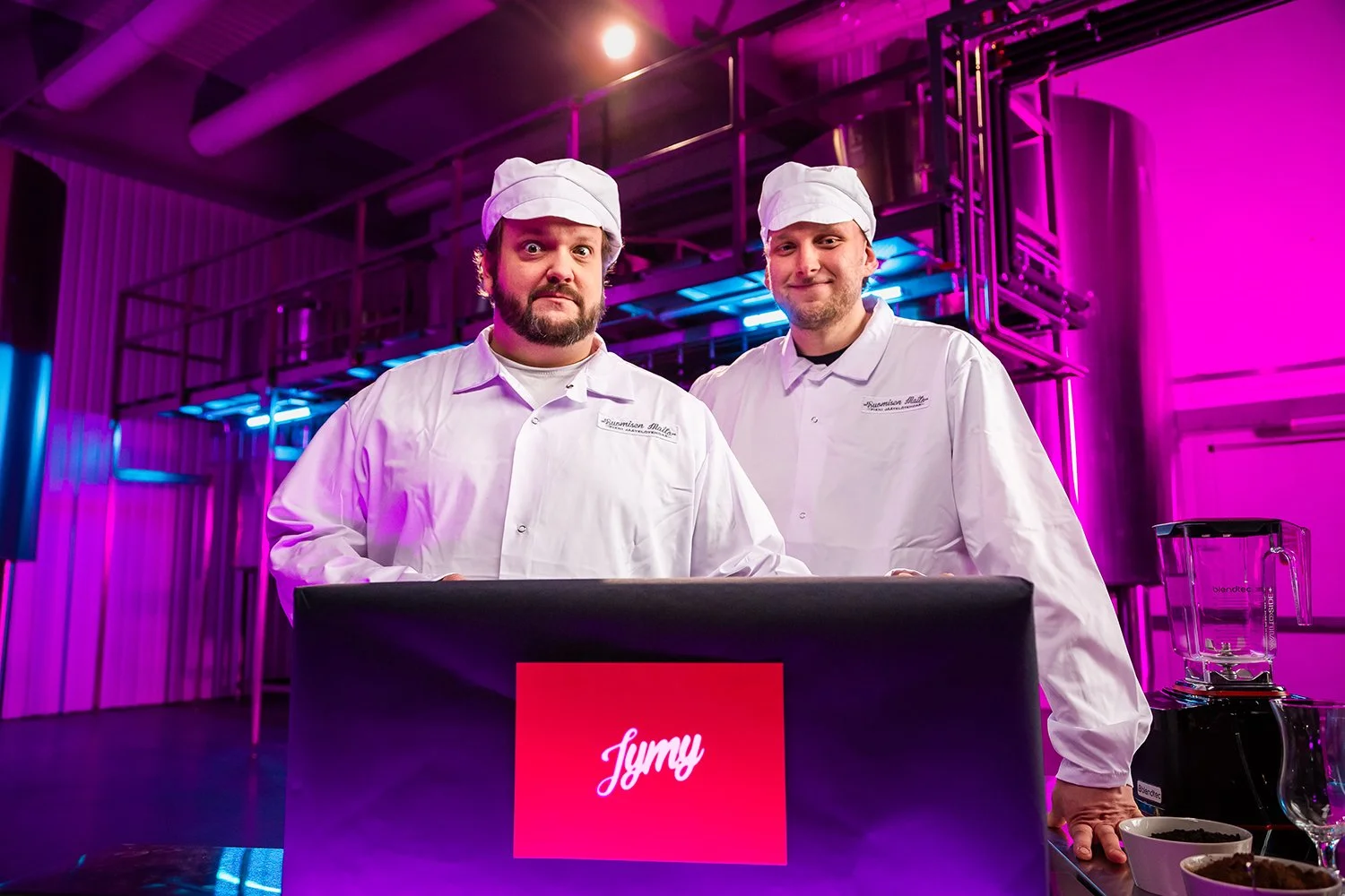 Kaksi miestä pukeutuneena kokin asuun, valkoisiin takkeihin ja hattuihin, seisovat modernissa keittiössä valaistuna violetein ja sinisin valoilla. He ovat edessä musta pöytälevy, jossa on punainen tulostemerkintä 'Jymy'. Taustalla näkyy keittolaittei
