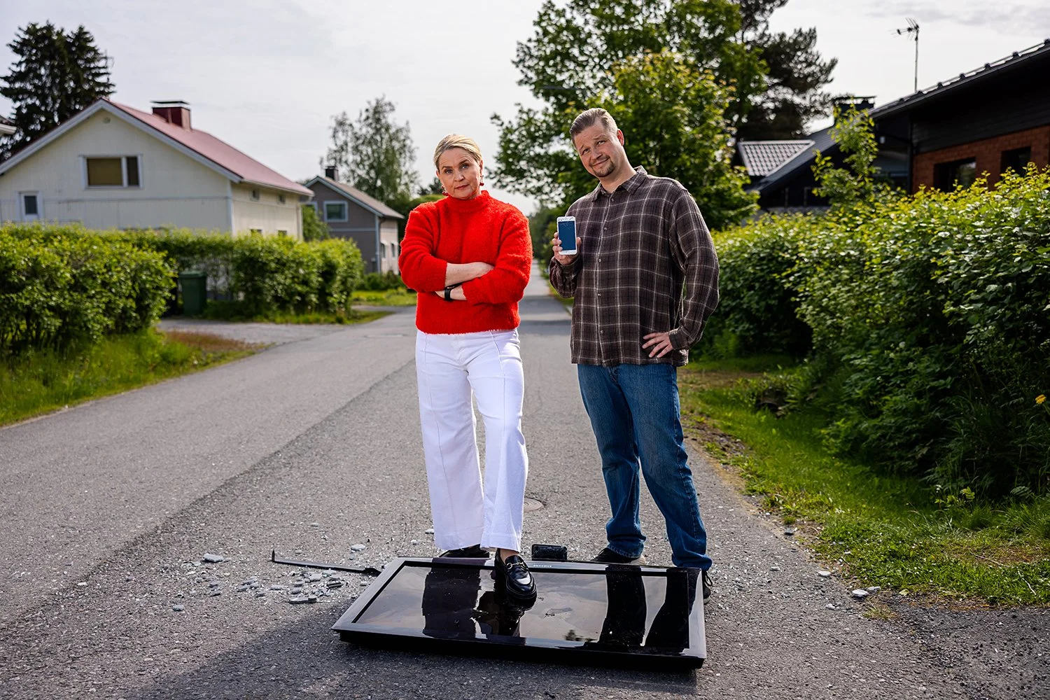 Kaksi henkilöä seisoo keskellä katua, toinen nainen punaisessa neuleessa ja valkoisissa houseissa, mies puku päällä, yksi heistä näyttää olevan varustautunut murtamaan ajoneuvon, toinen pitelee puhelintaan.