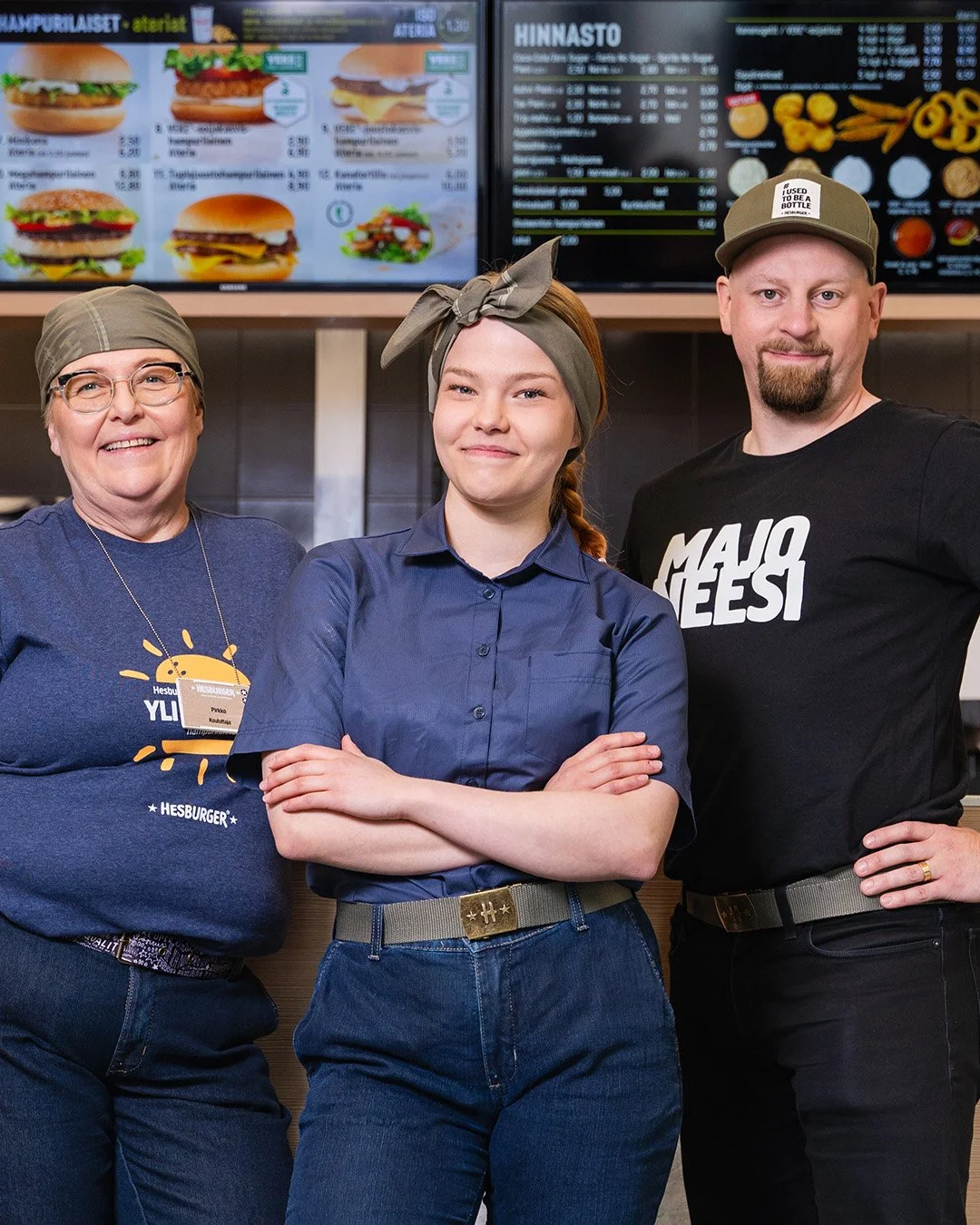 Kolme ihmistä poseeraamassa hampurilaisravintolassa, taustalla ruokalistat näytöillä.