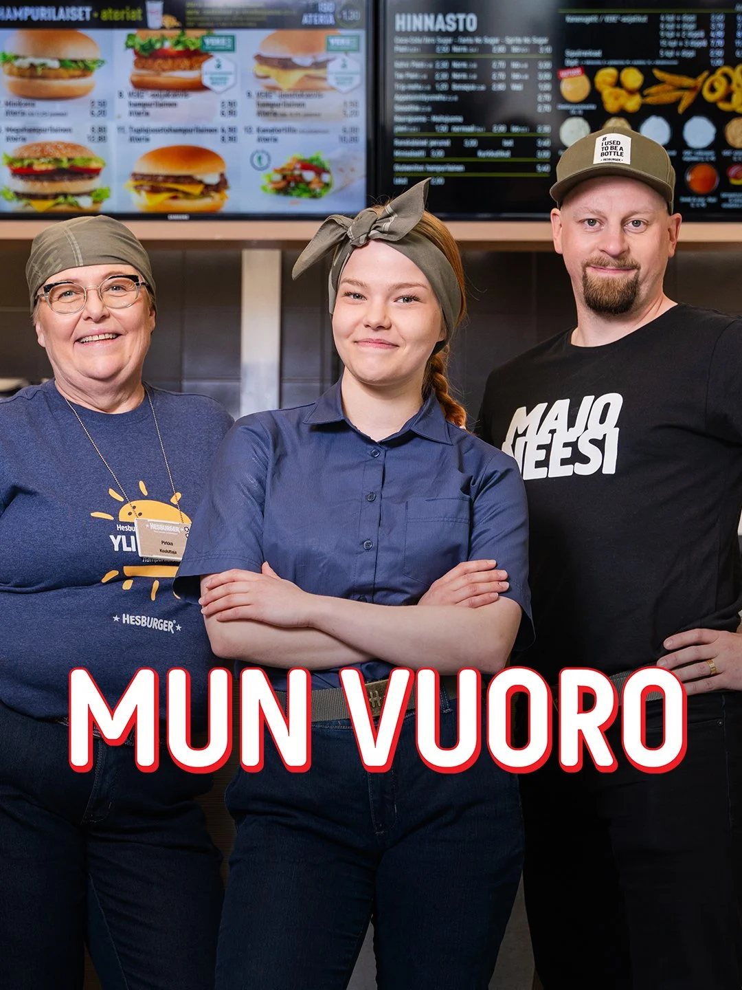Kolme ihmistä seisoo ravintolassa, taustalla ruokalista näytöillä ja tekstillä "MUN VUORO".