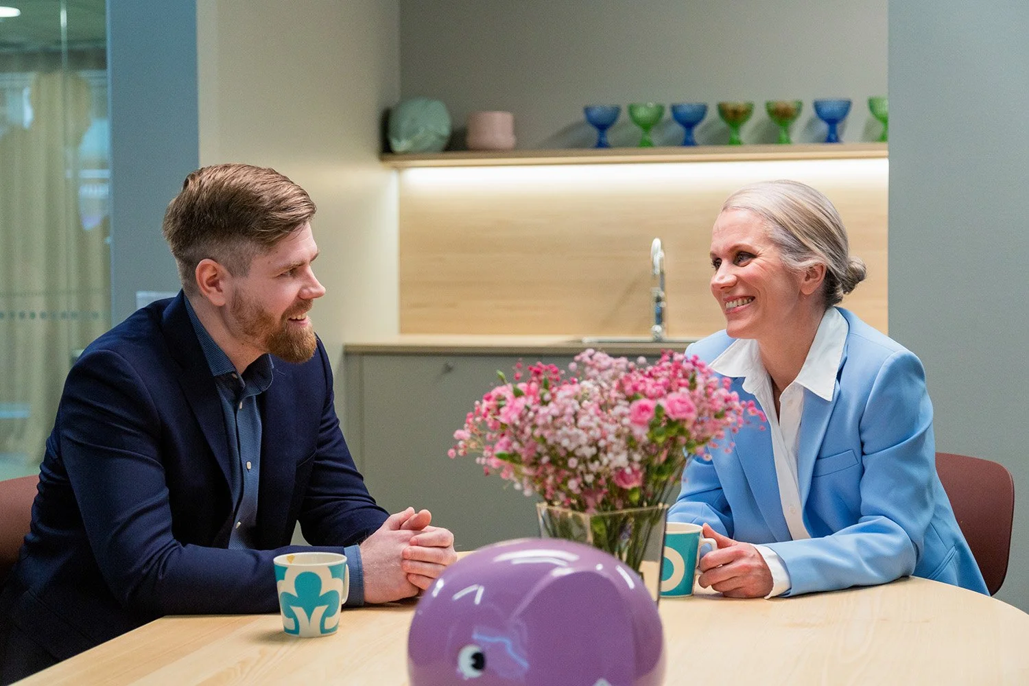 Kaksi aikuista istuu pöydän ääressä, keskustellen ja hymyillen, kahvikupit kädessä. Pöydällä on suuri pinkki kukkakimppu ja lelu-ankka.