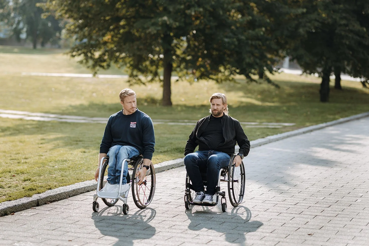 Kaksi miestä pyörätuoleilla kävelee puiston polulla vuorotellen ja keskustelevat.
