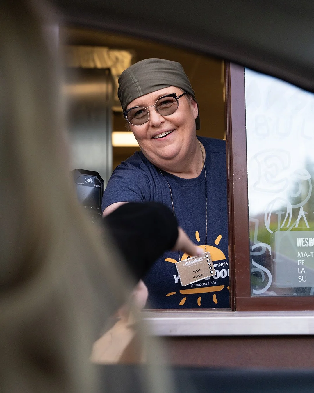 Hesburgerin myyjä palvelee asiakasta ikkunassa, hänellä on päässä harmaa hattu, silmälasit ja Sininen Hesburger T-paita.