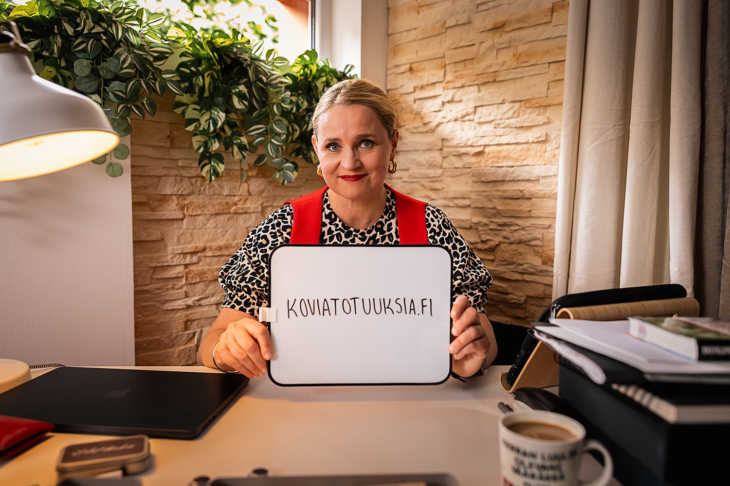 Nainen istuu työpöydän ääressä, pitää paperilautasta, jossa lukee 'KOVATOTUUKSIA.FI', ja hymyilee. Taustalla kivi- ja verhoikkuna, pöydällä läppäri ja kirjoja, ja pöydän etualalla kahvimuki.