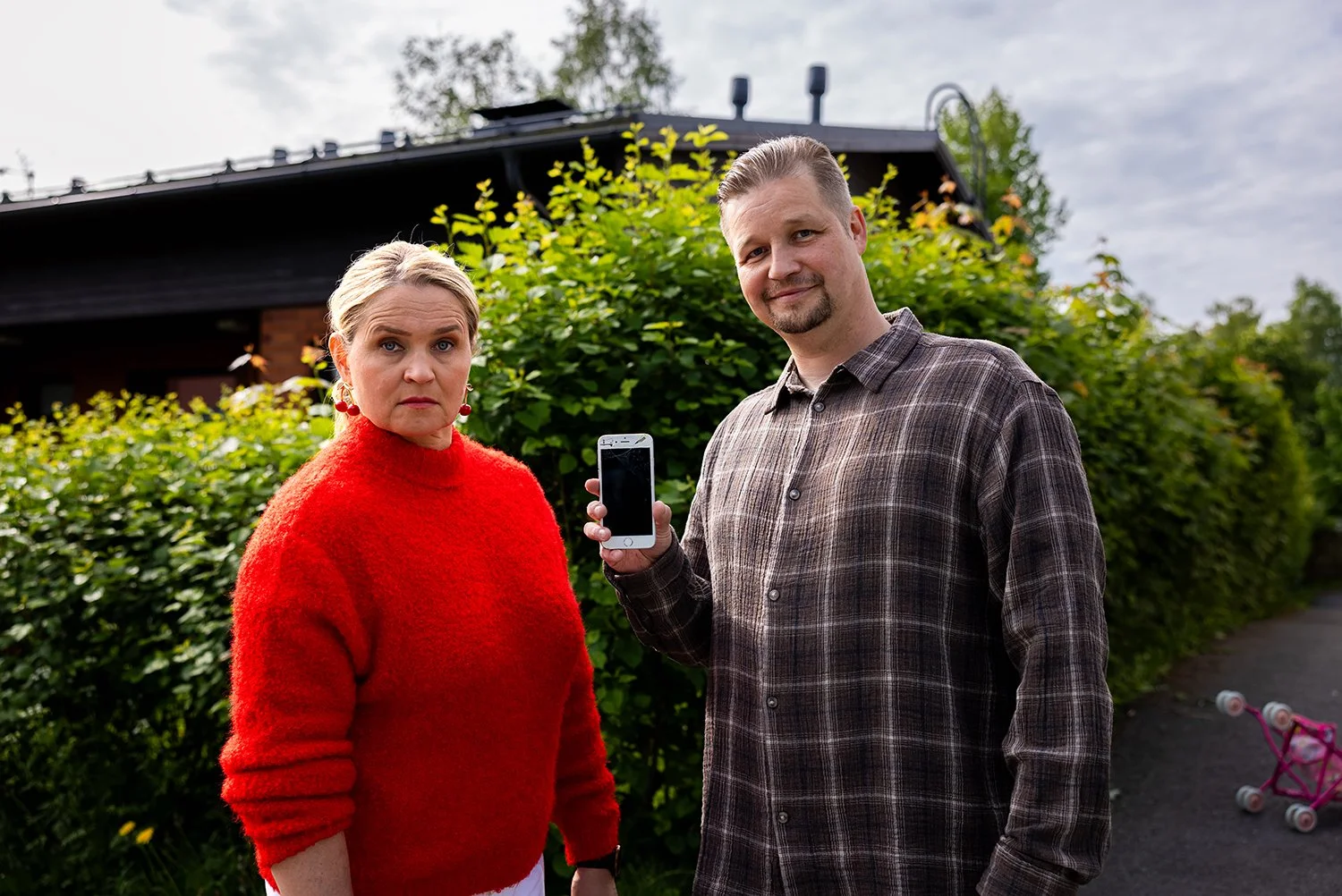 Kaksi ihmistä ulkona, nainen punaisessa neuleessa ja mies ruutupaidassa, mies näyttää kuvaan ja näyttää älypuhelinta, taustalla pensaita ja talo.