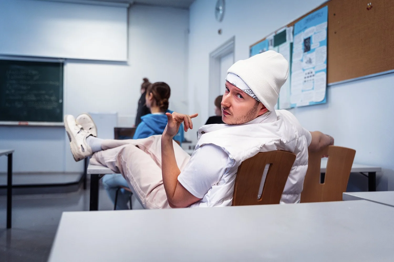 Nuori miesopiskelija istuu luokkahuoneessa, katsoo kameraan ja tekee tyylikkään ilmeen, hänellä on valkoinen pipo ja valkoinen pusero. Taustalla näkyy muita opiskelijoita ja ilmainen musta taulu.