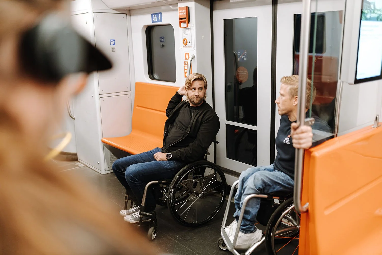 Kaksi nuorta miestä istuu ratikkavaunussa, toinen istuu pyörätuolissa ja toinen on istumassa. Kolmas henkilö, jonka kasvot ovat osittain näkyvissä, on lähietäisyydellä vasemmalla puolelta. He ovat matkalla kaupungissa.