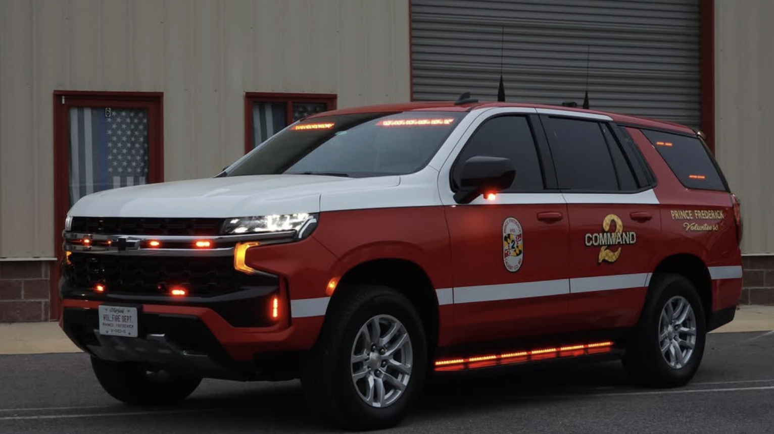 A red and white emergency response vehicle with 'Prince Frederick Volunteer' markings and lights on, parked in front of a beige building.