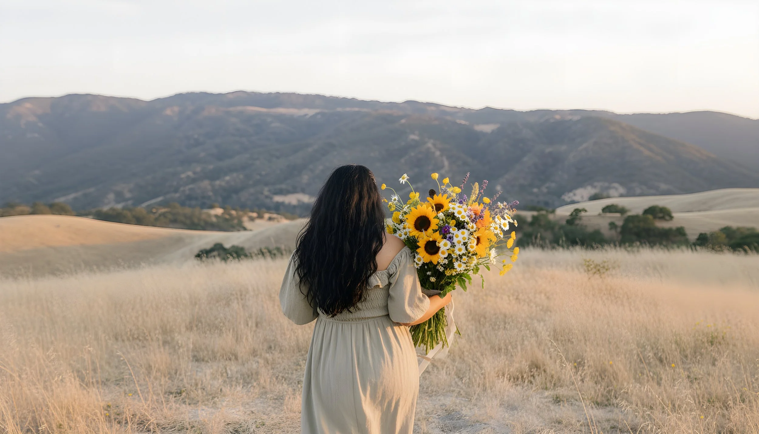 A woman with long dark hair wearing a light-colored dress holding a large bouquet of sunflowers and other wildflowers in a field with rolling hills and mountains in the background.