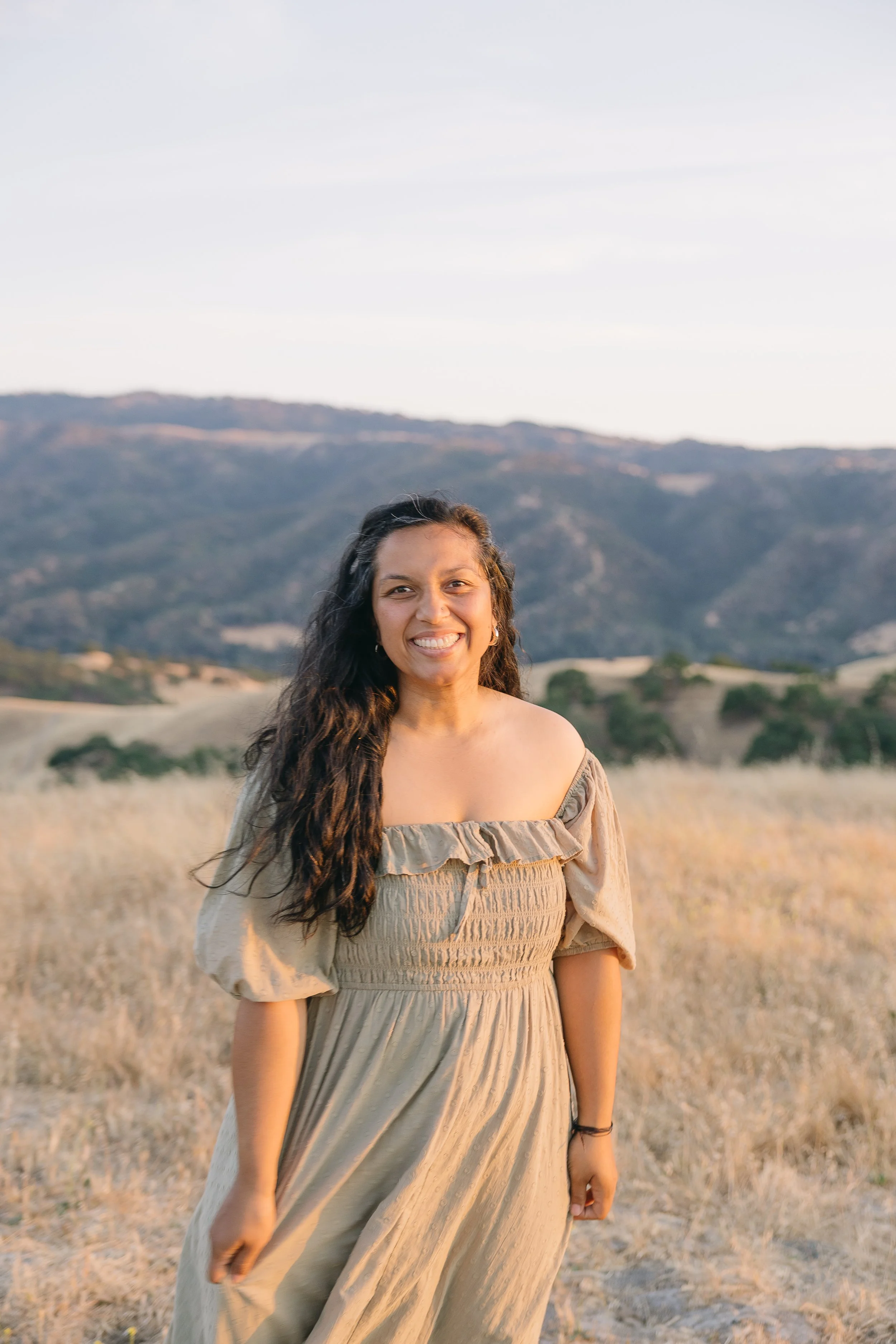 A woman with long, dark, curly hair smiling at the camera in a beige dress in a field with mountains in the background during sunset.