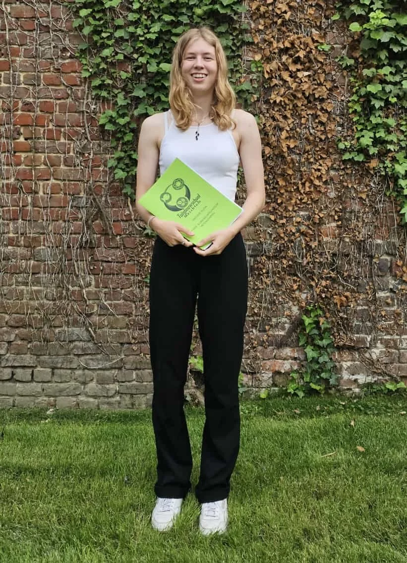 Een jonge vrouw met lang, golvend blond haar staat op een grasveld voor een bakstenen muur die gedeeltelijk bedekt is met groene en bruine klimop. Ze draagt een witte tanktop, zwarte broeken en witte sneakers. Ze houdt een groene map vast en lacht naar de camera.