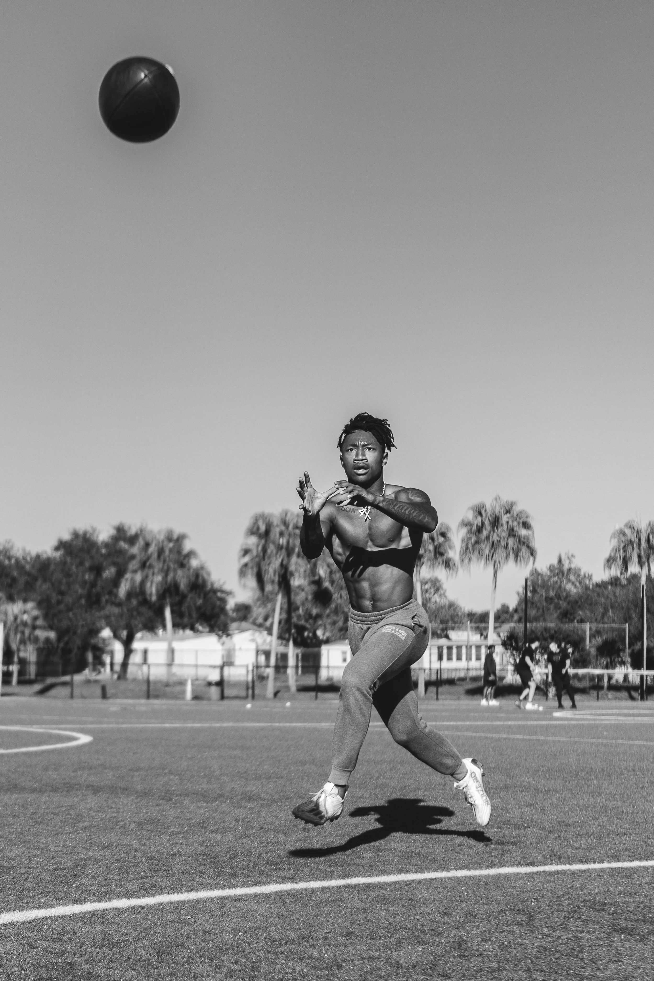 A shirtless young man wearing sweatpants and sneakers playing football on an outdoor court, jumping to catch or intercept the ball, with a clear sky and palm trees in the background.